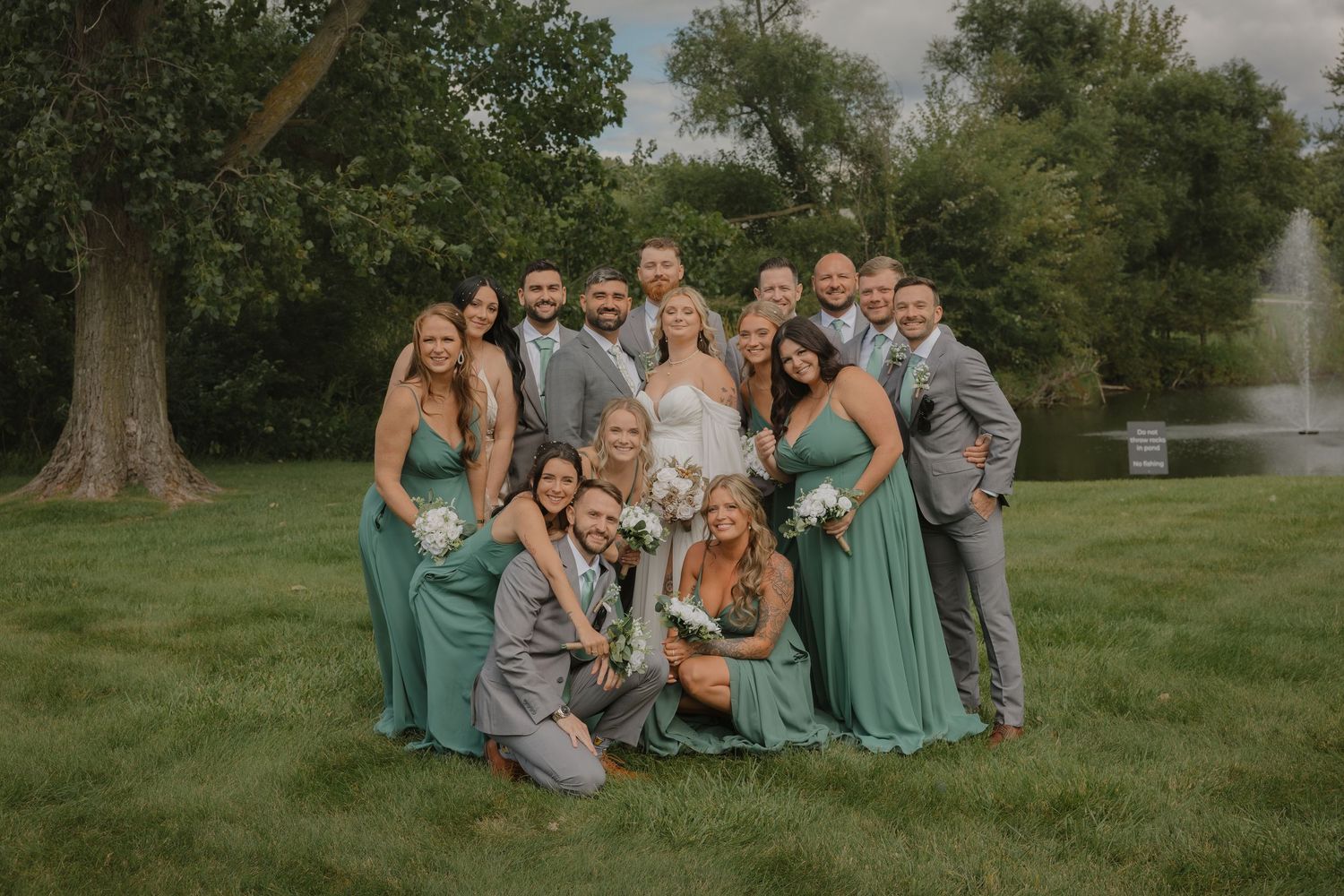 Wedding party members pose casually on grass in emerald and gray attire.