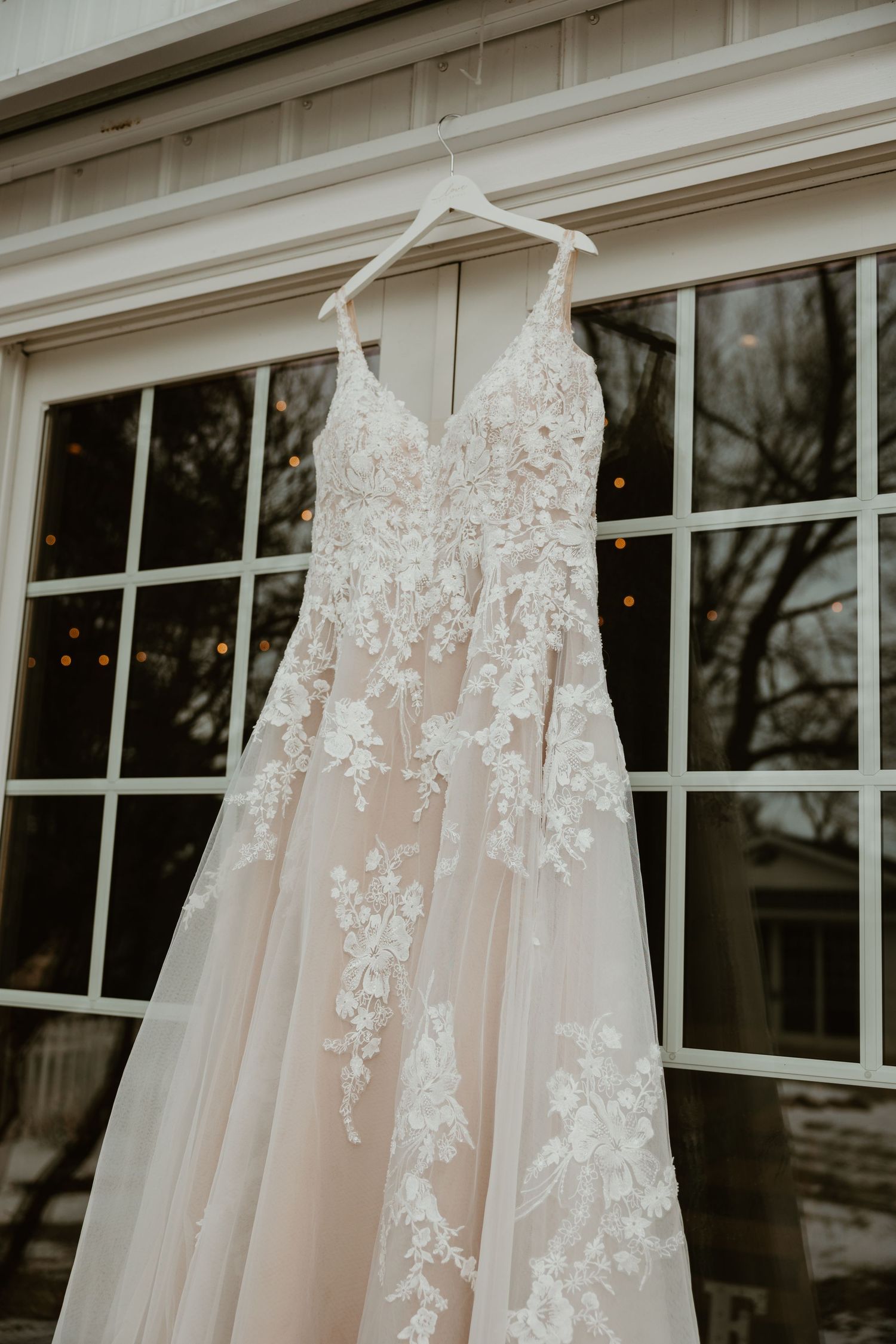 A detailed shot of a blush wedding gown with lace appliques hanging against a window.