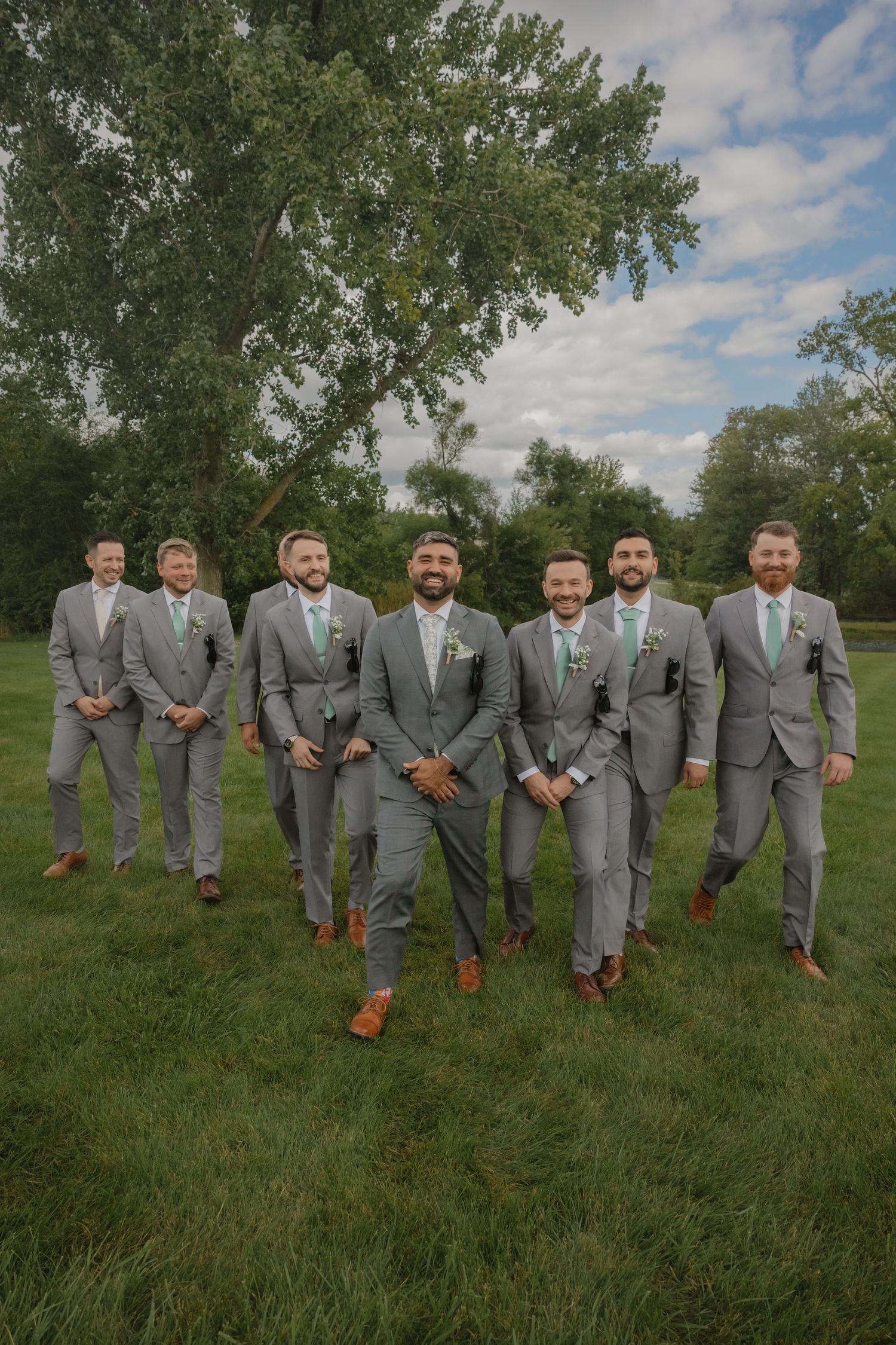 Groomsmen in gray suits with brown shoes pose playfully on grass for wedding photos.