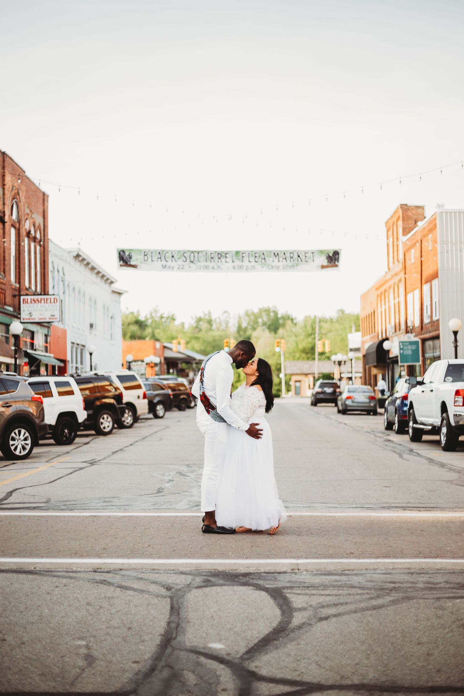 A couple in wedding attire embraces in the middle of a small town street lined with brick buildings and parked cars.