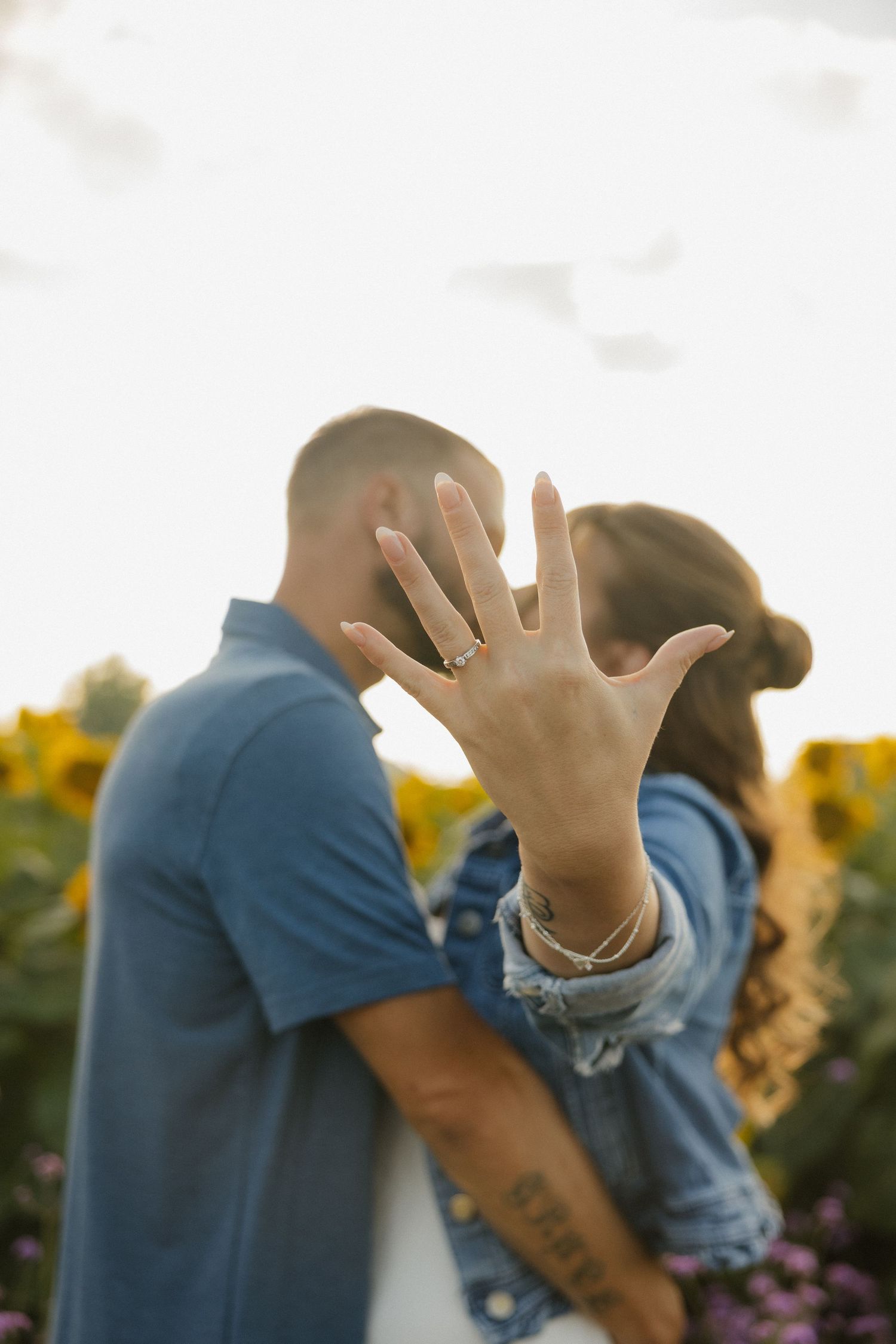 Person in blue shirt playfully covering face and gesturing with hands in front of sunflower field at sunset.
