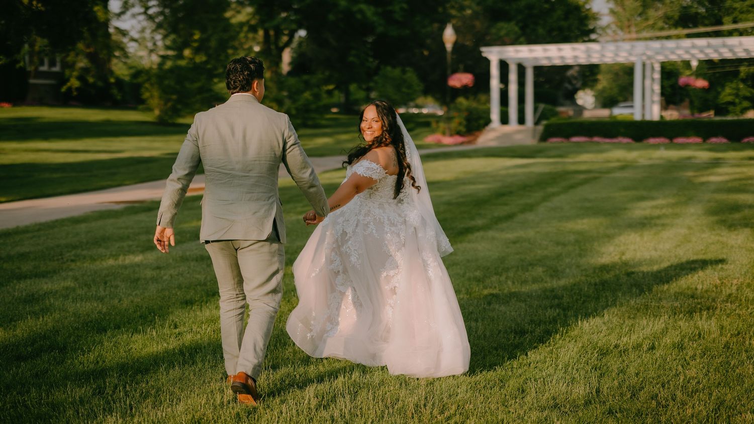 A couple in wedding attire walking together on grass in golden light at sunset.