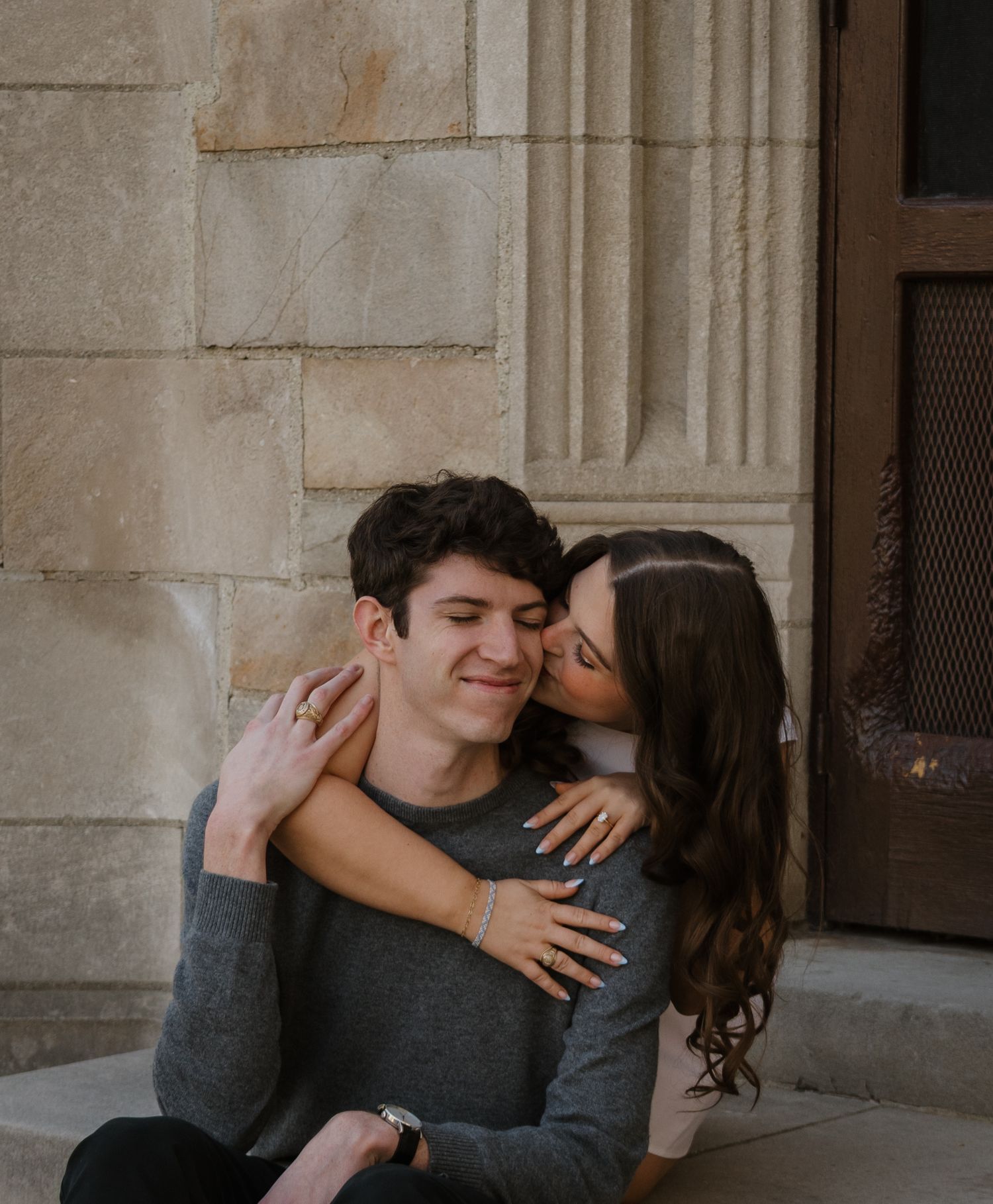 A couple sits on stone steps, woman kissing man's cheek as he smiles, both wearing gray sweaters.