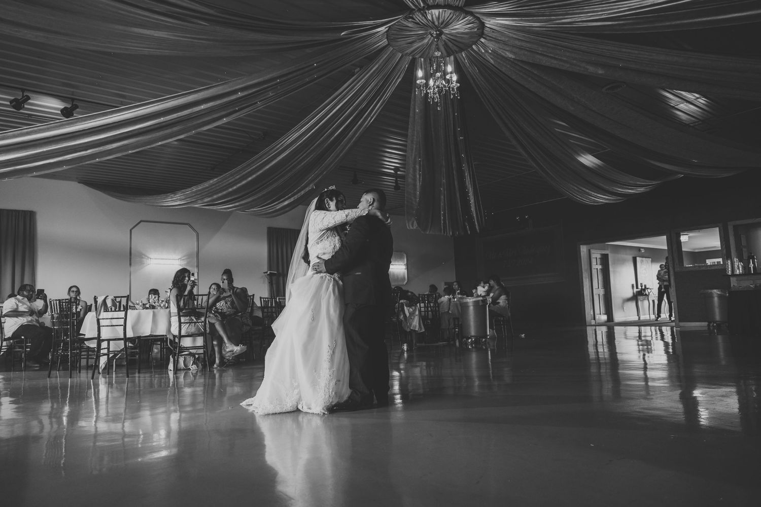 Black and white photo of a couple sharing their first dance under elegant draped fabric and chandelier.