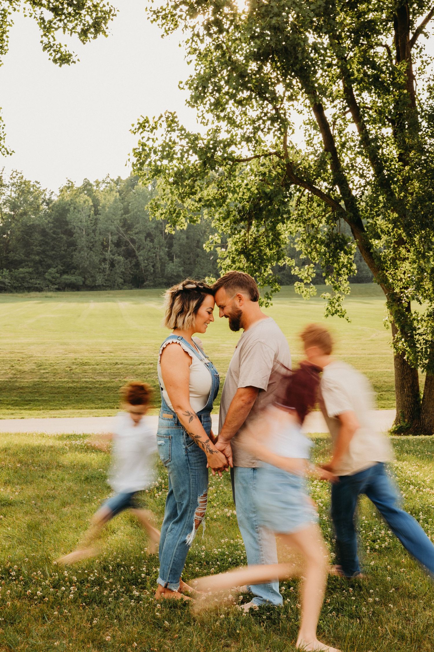 Couple shares intimate moment while others run playfully in background under tree canopy.