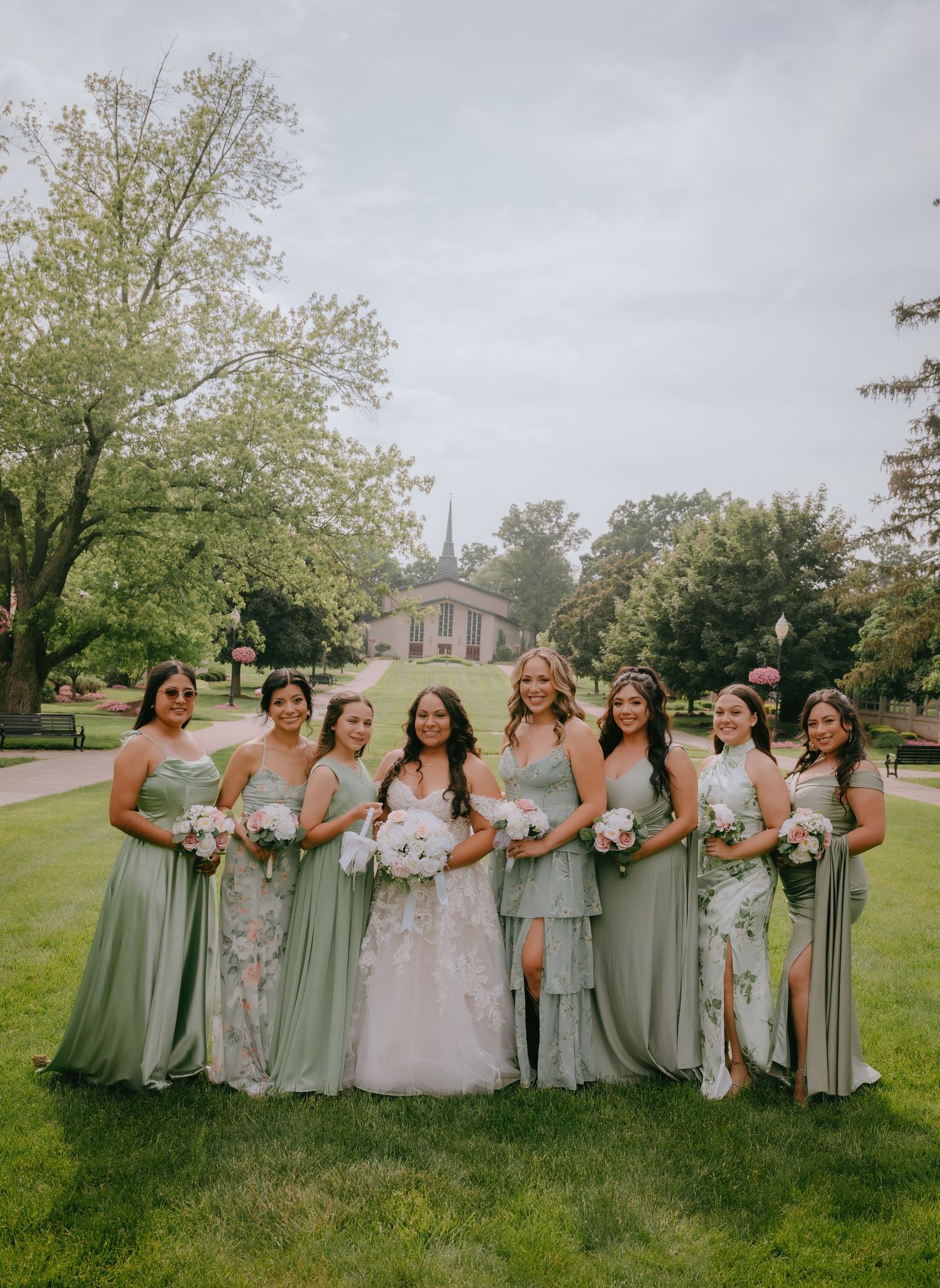 Wedding party in sage green bridesmaid dresses poses on a lush lawn with trees and a chapel in the background.