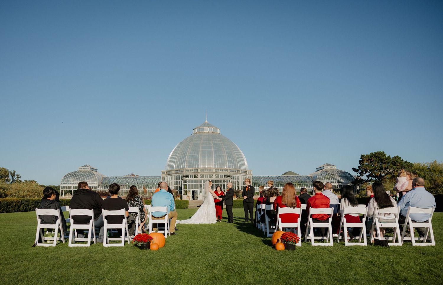 Outdoor wedding ceremony on green lawn before grand glass conservatory dome under clear blue sky.