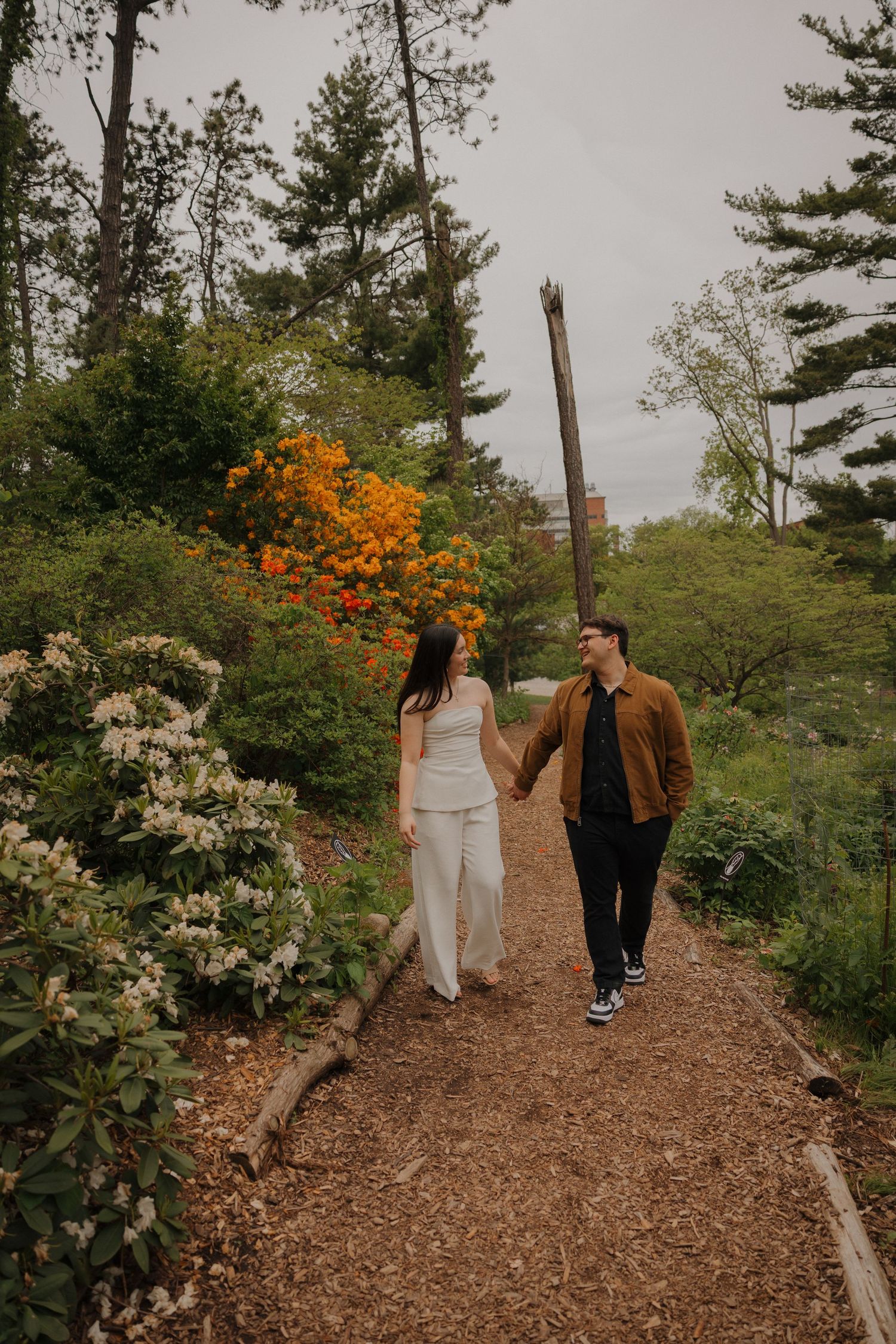 Couple holding hands walking on a garden path surrounded by colorful spring blooms and tall evergreen trees.