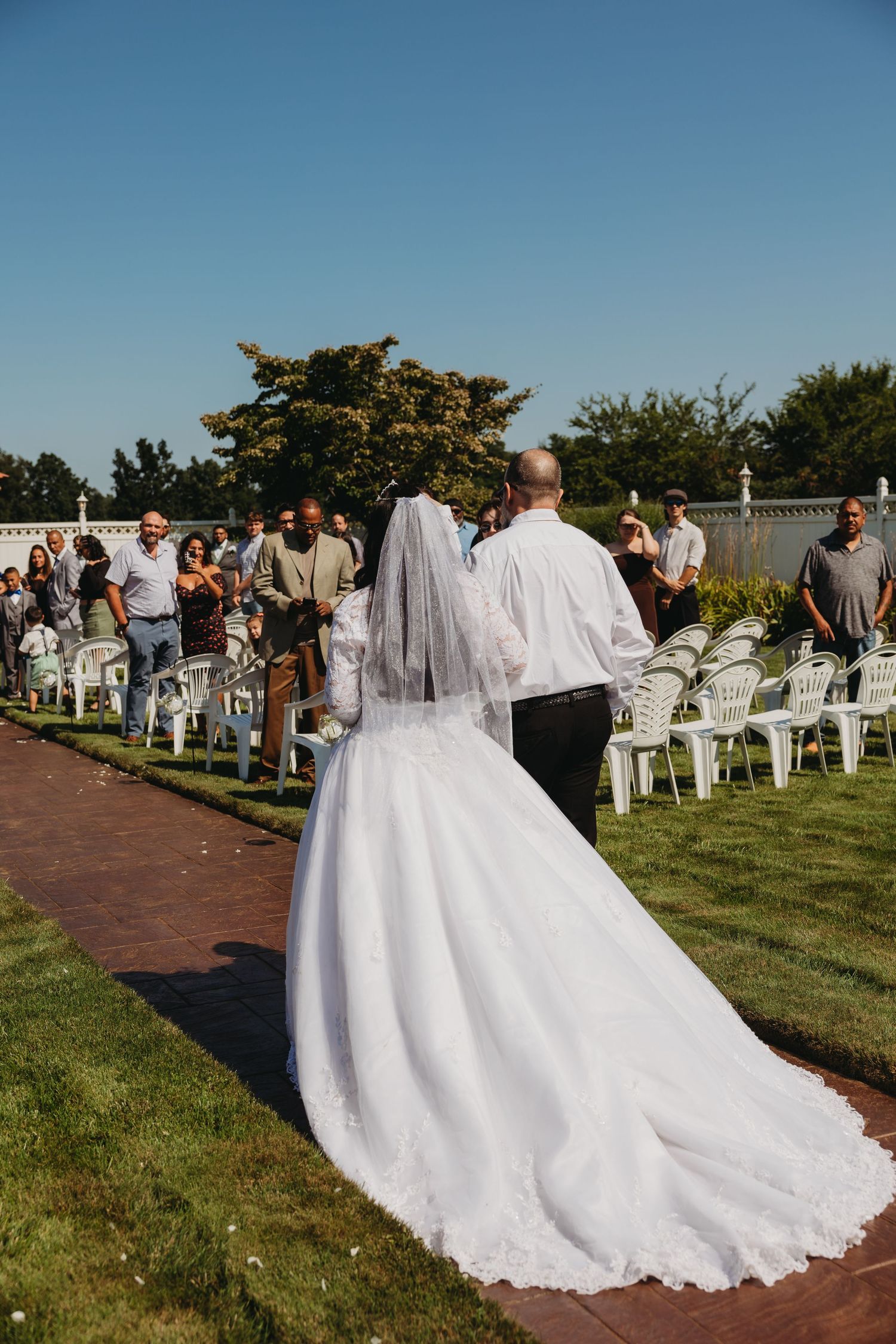 Long white wedding dress train flows down outdoor aisle with guests seated on white chairs.