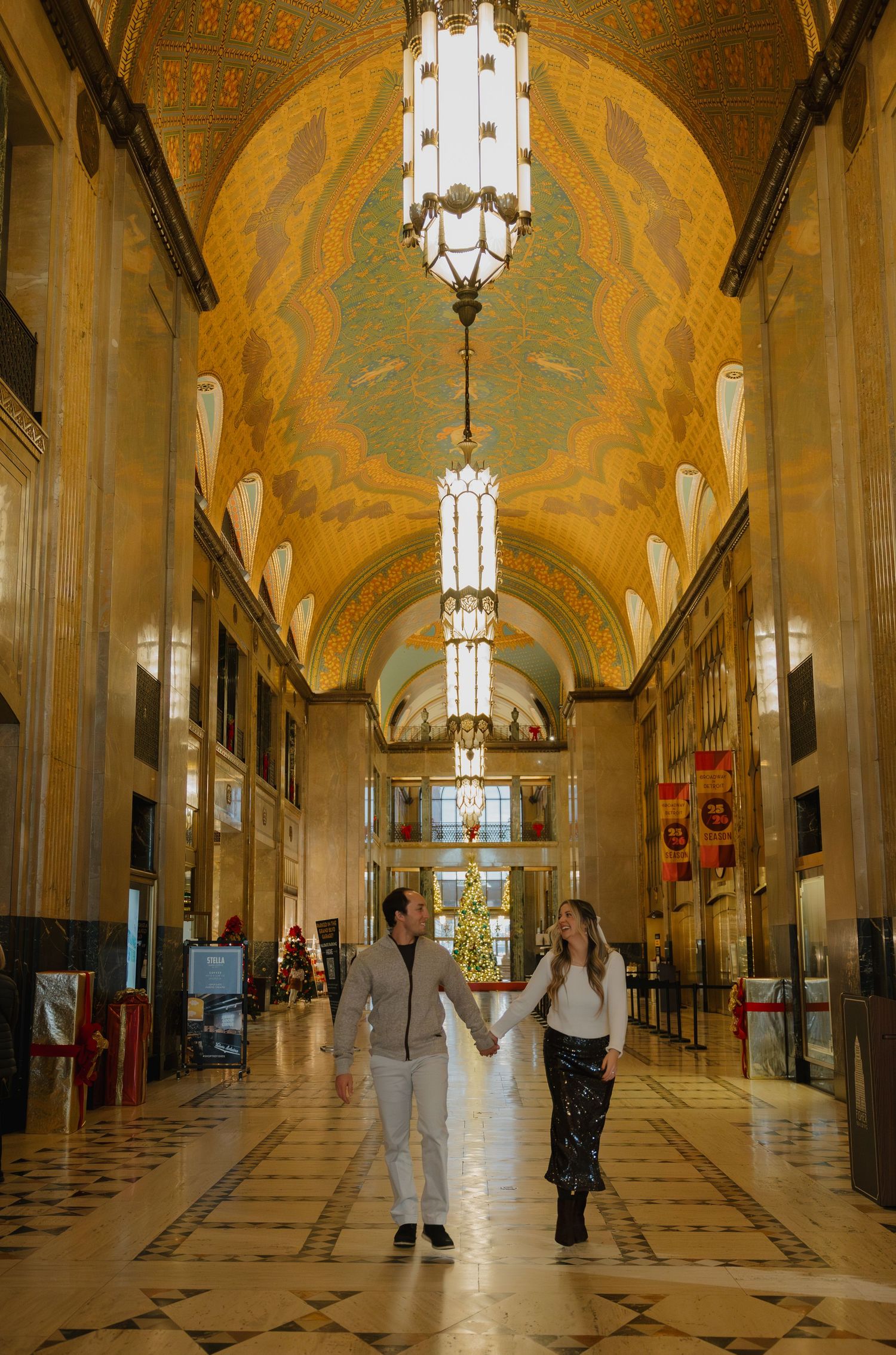 Couple holding hands walking through ornate Art Deco building lobby with gilded arched ceiling and elegant chandeliers.