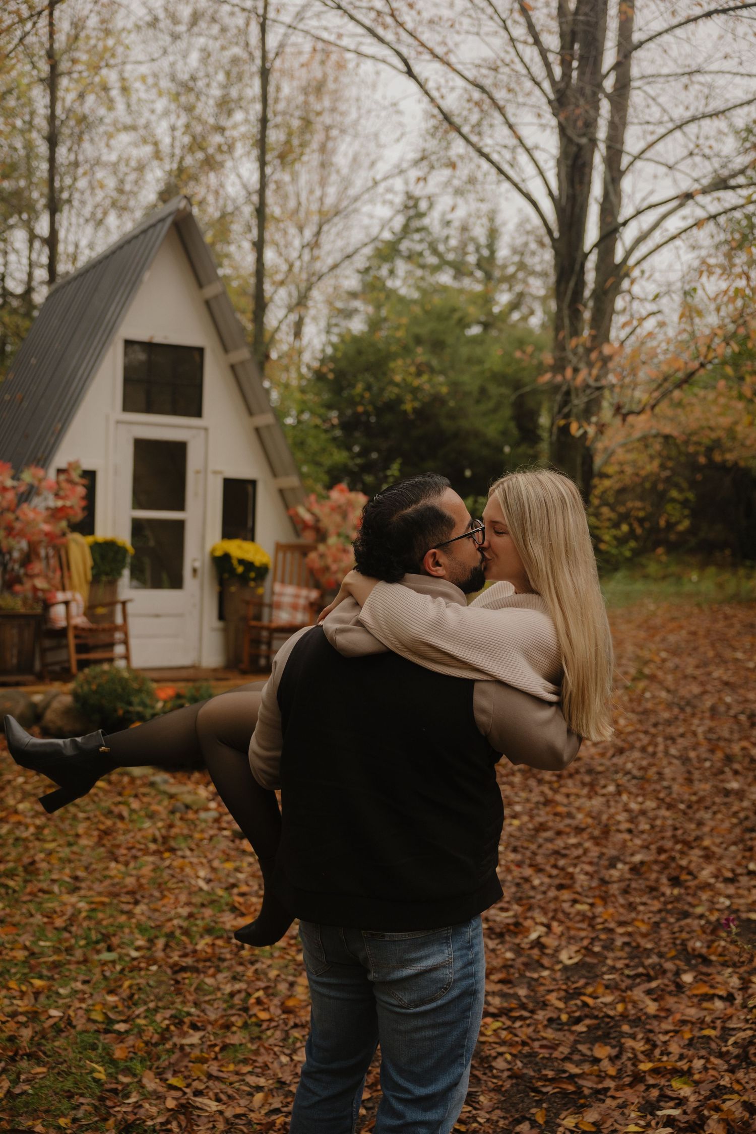 A couple dances together in fallen autumn leaves outside a white A-frame cabin in the woods.