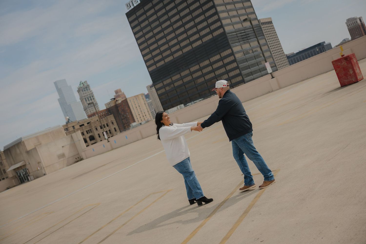 Couple dancing on rooftop with city skyline backdrop, man in cap and jeans, woman in white top and boots.