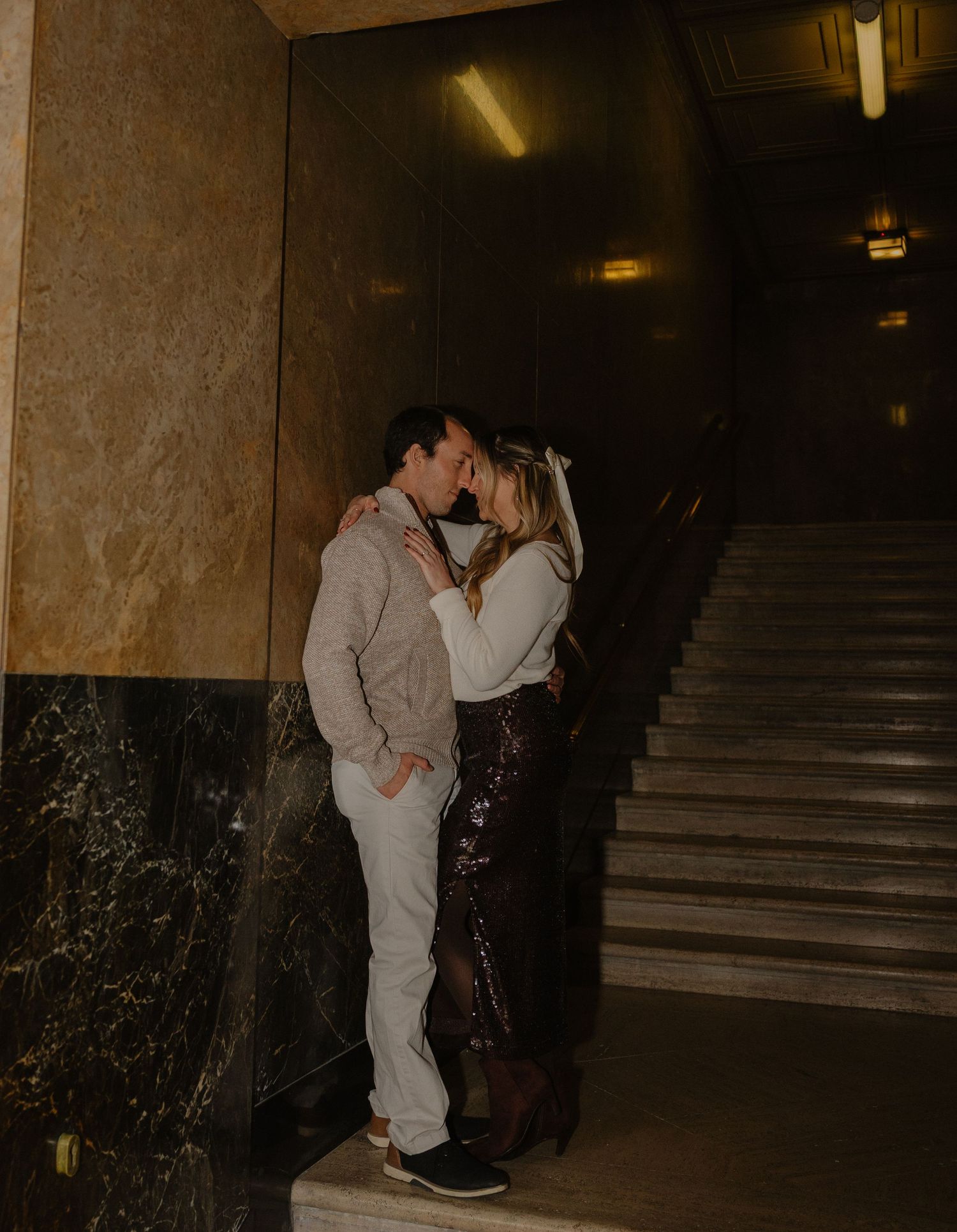 Couple sharing a romantic moment near marble stairs in a dimly lit building interior.