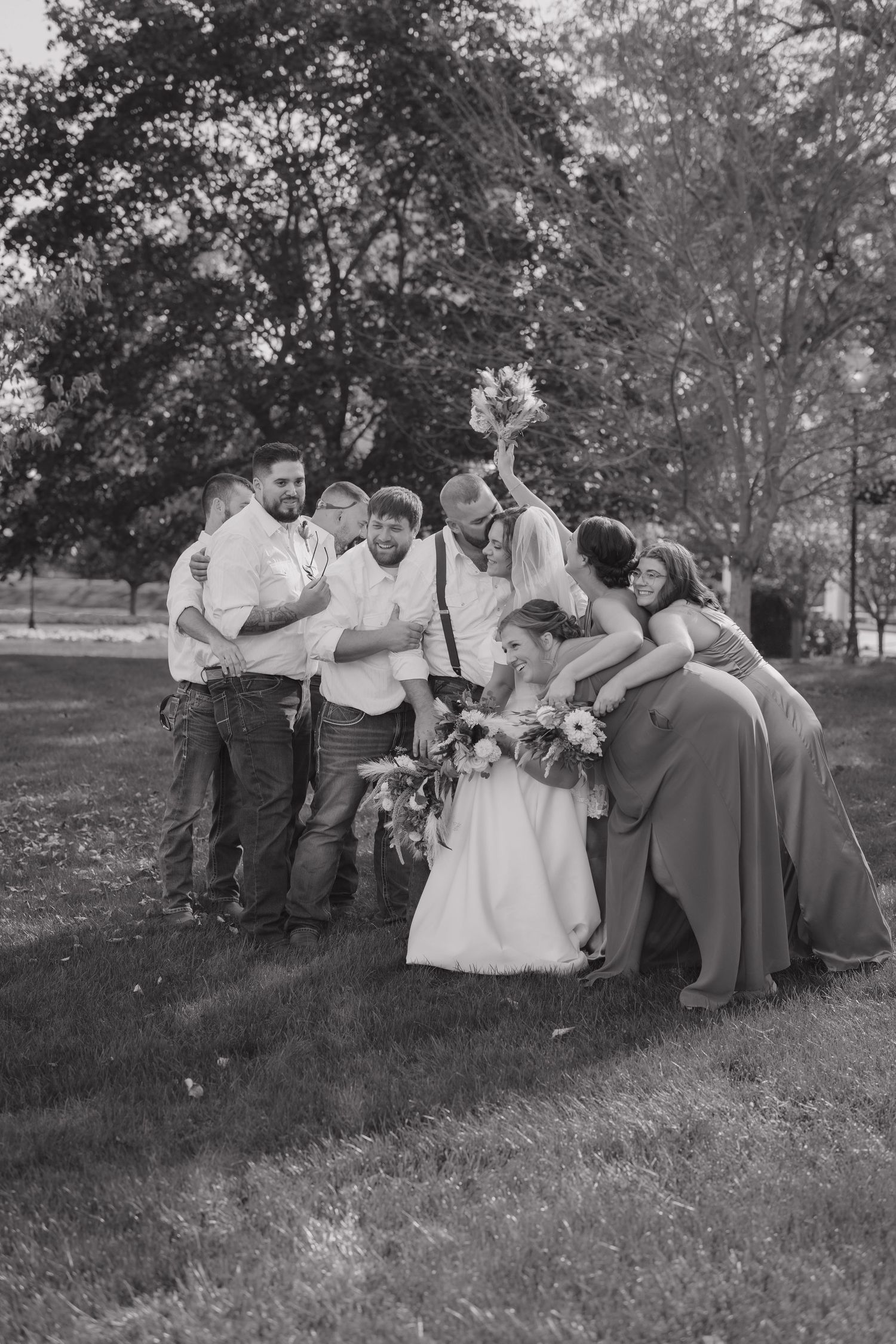 Black and white wedding photo of bride, groom, and bridal party celebrating joyfully outdoors on a grassy lawn.