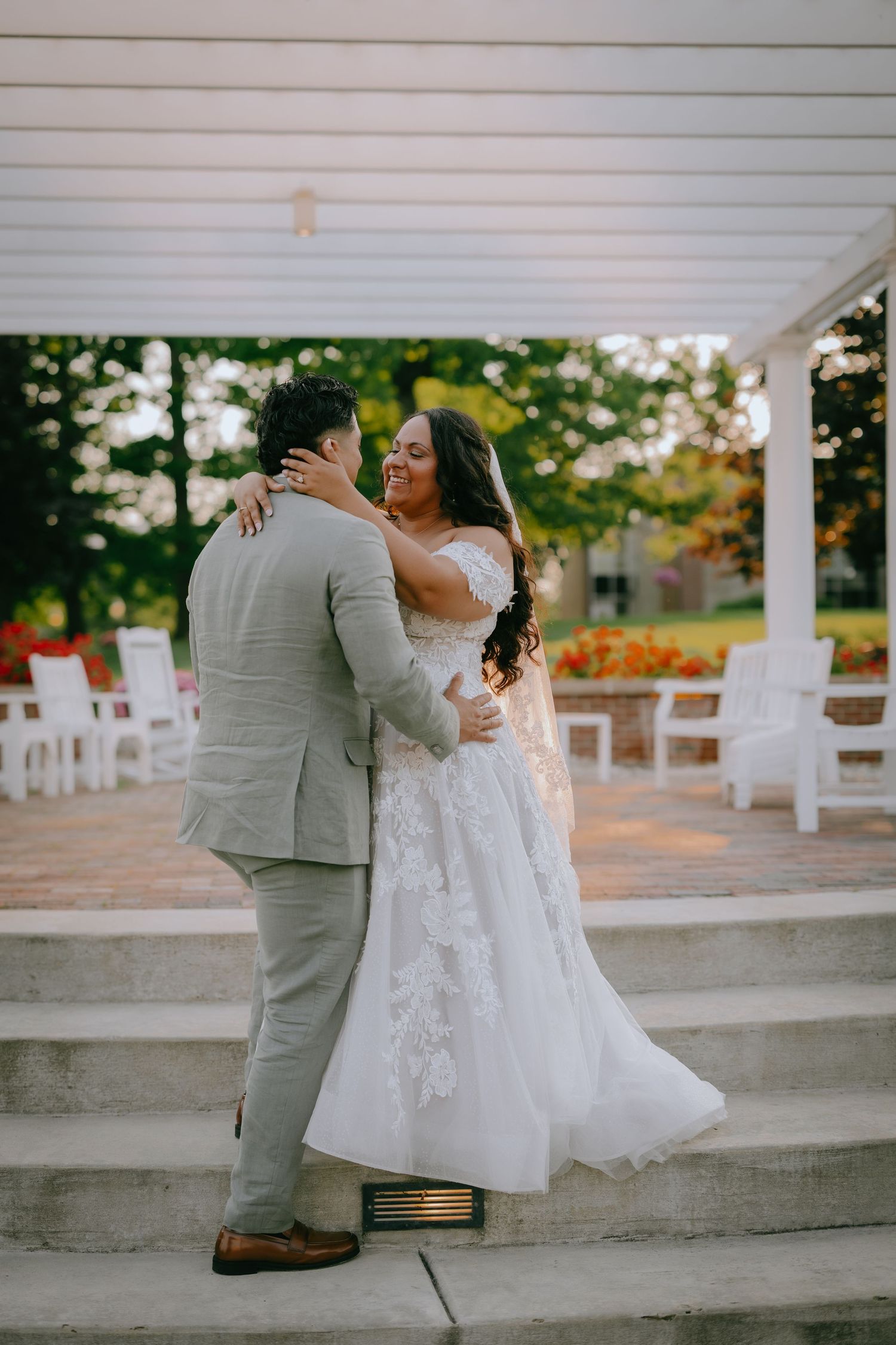 Newlywed couple shares romantic dance moment on outdoor patio steps with white chairs and greenery in background.