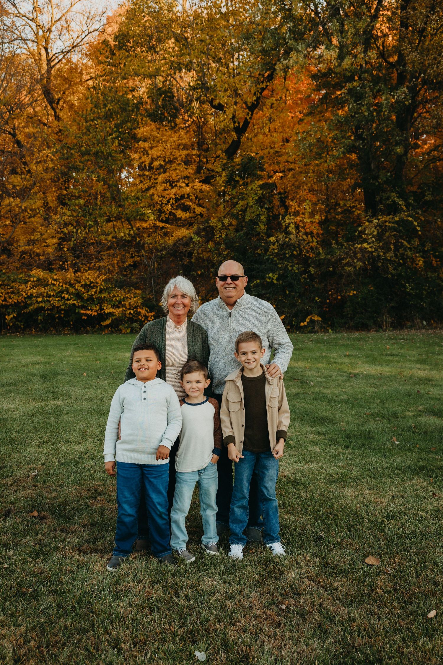Grandparents pose with three grandchildren against vibrant fall trees in outdoor setting.