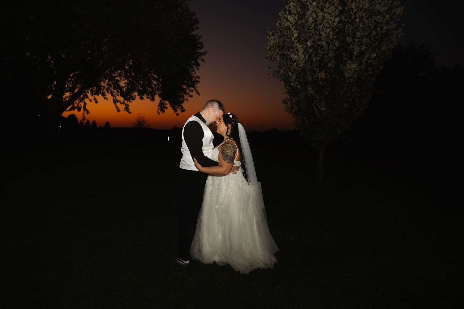 Silhouettes of a couple in wedding attire embrace during a dramatic sunset behind tall trees.