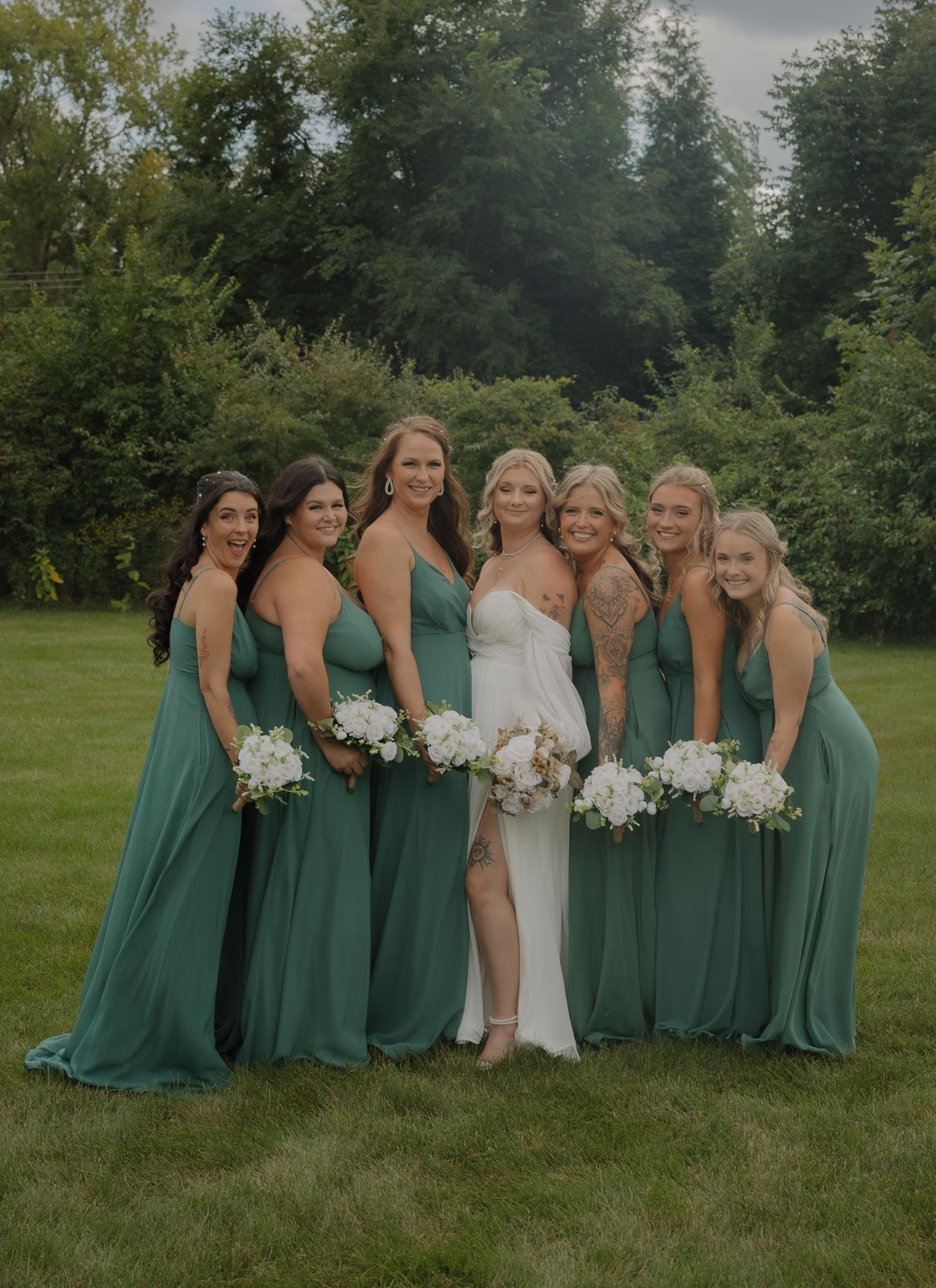 Wedding party in emerald green bridesmaid dresses posing together on grassy field.