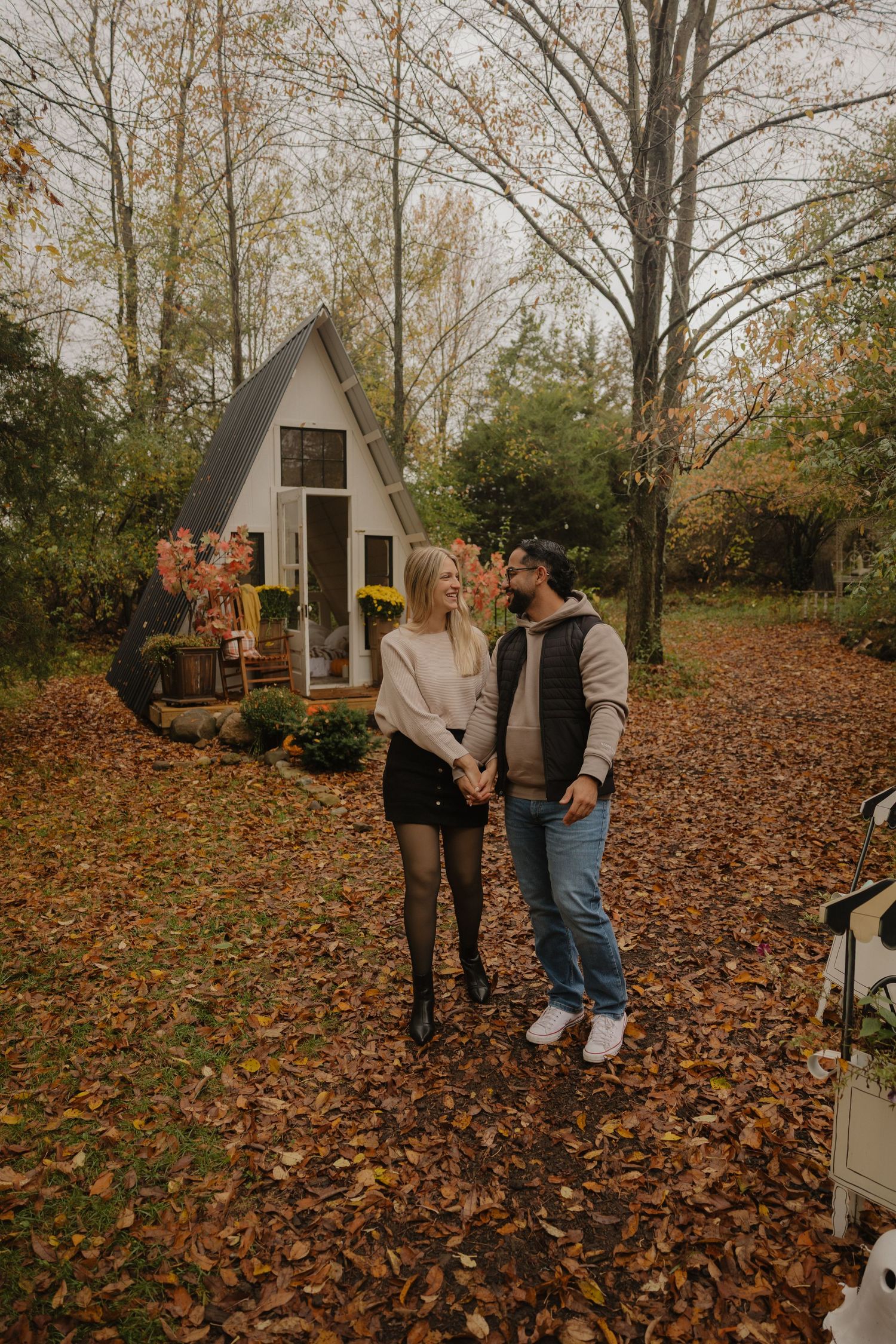 A couple enjoys a romantic autumn moment among fallen leaves near a small A-frame cabin in the woods.