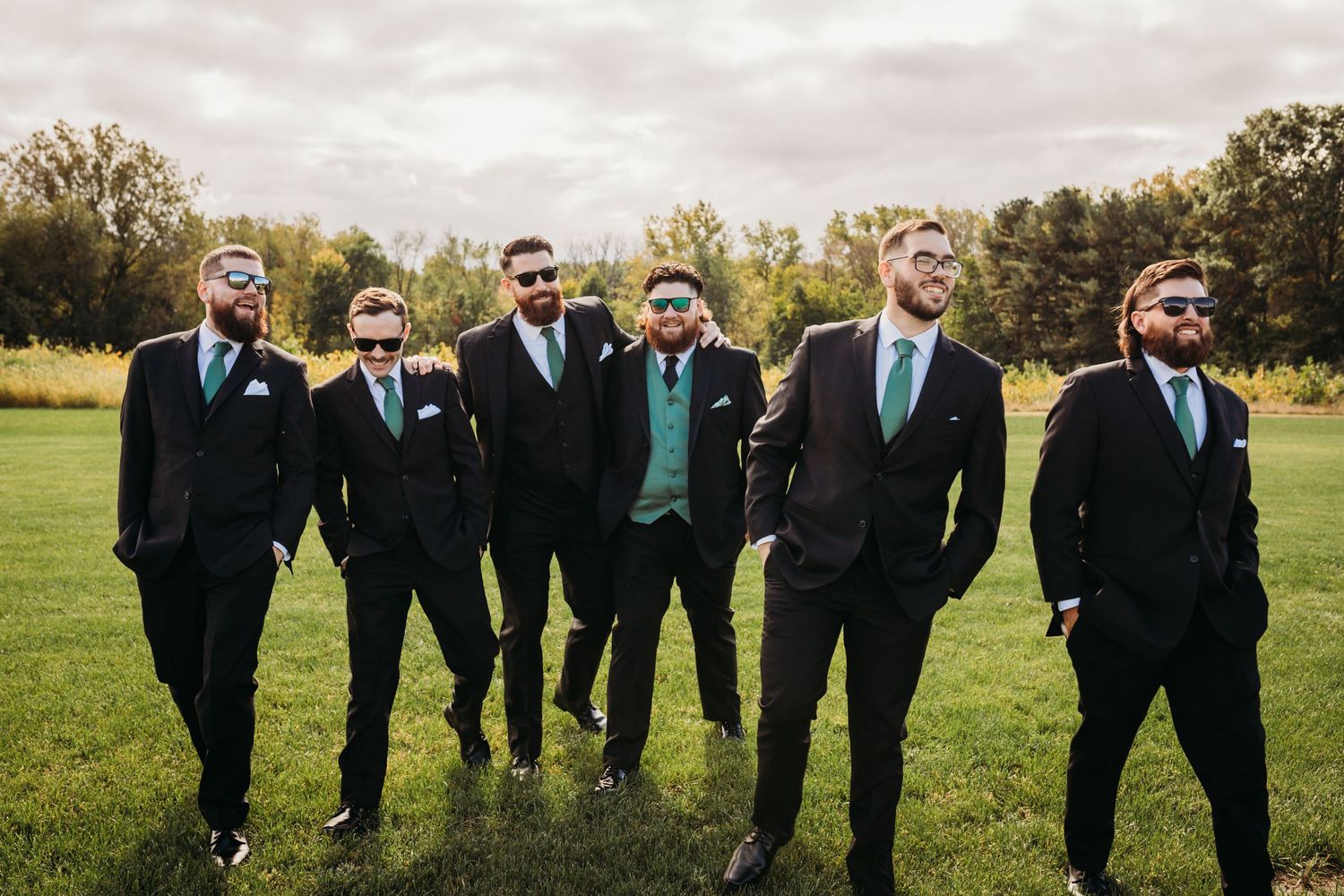 Groomsmen in black suits with teal accents pose confidently on grass field.