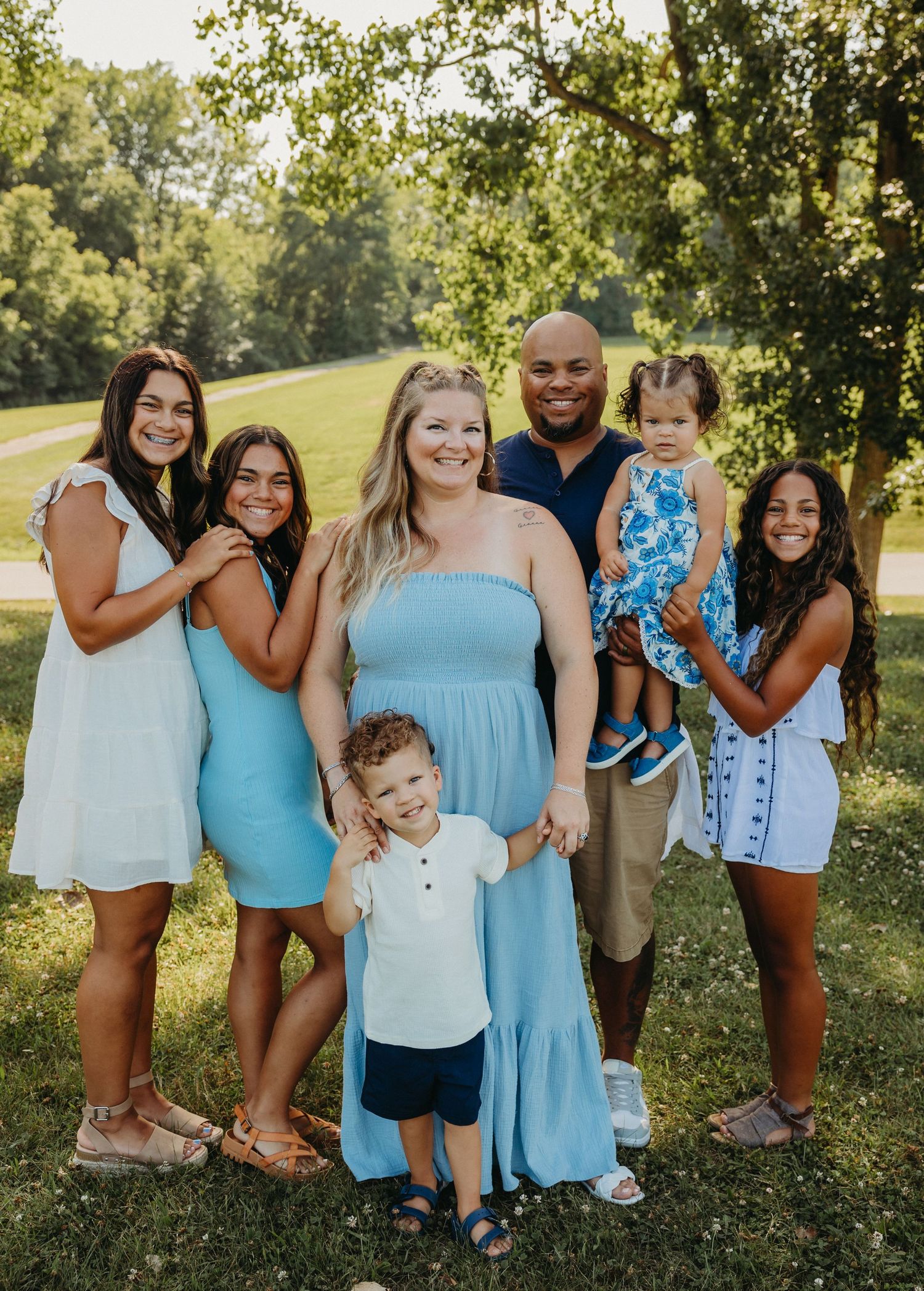 Family group portrait outdoors wearing coordinating blue dresses in a sunny park setting.