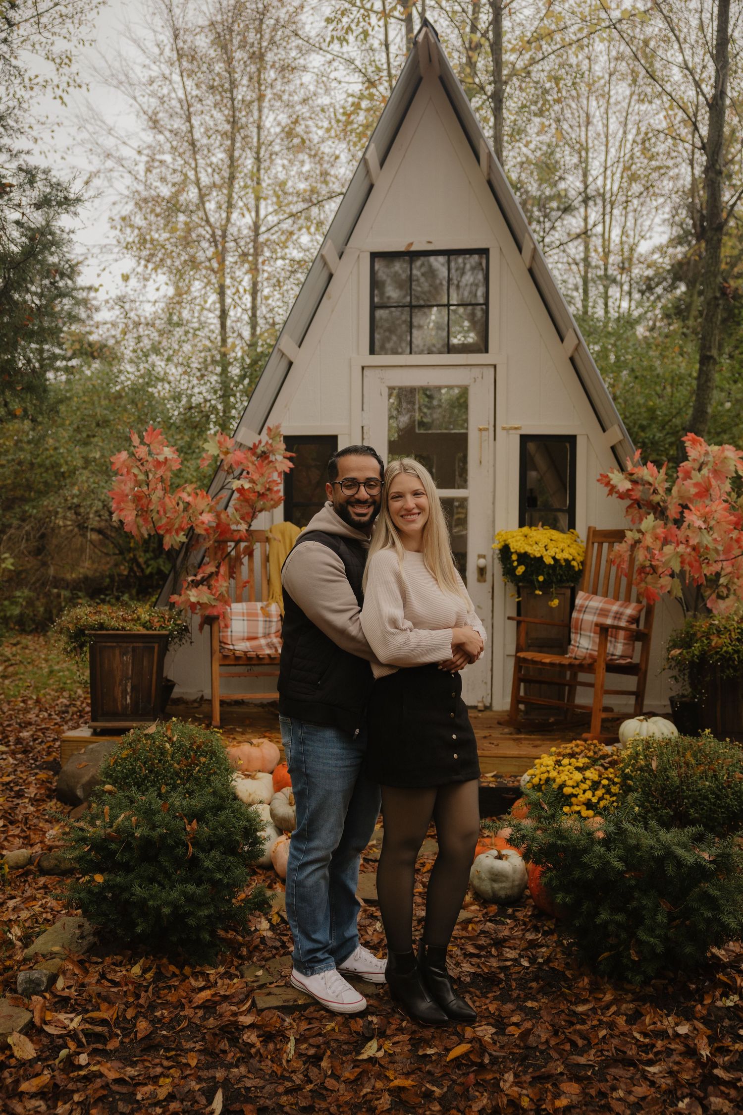 A couple stands in front of a cozy A-frame cabin surrounded by fall foliage and autumn decorations.