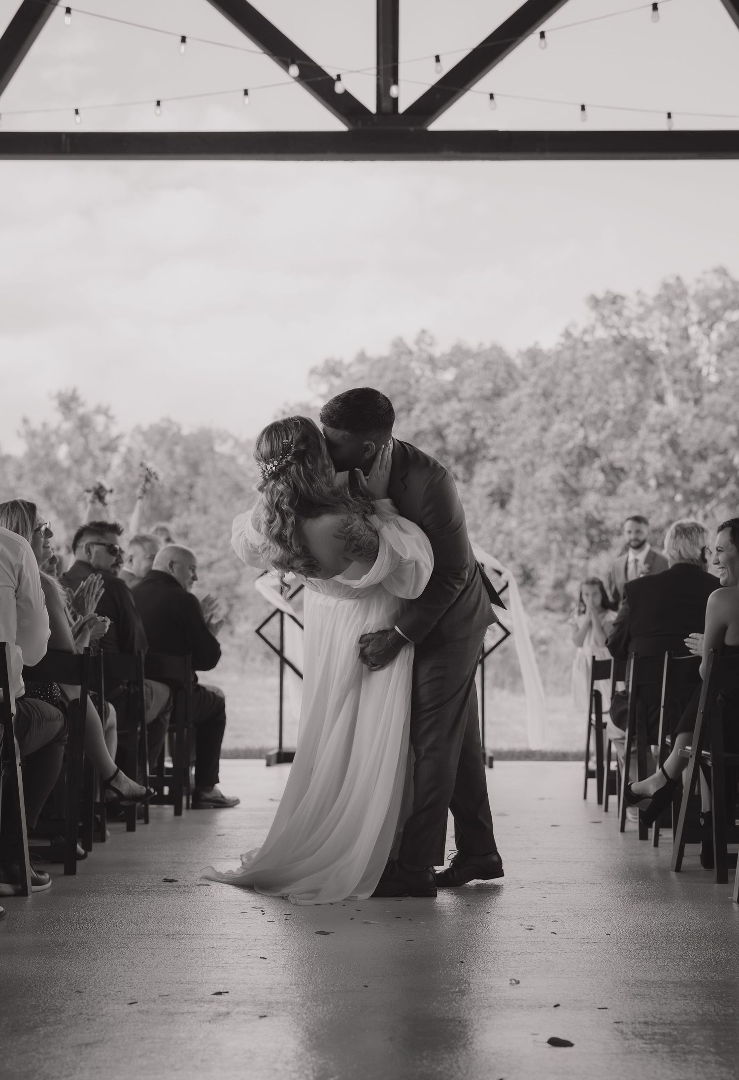 A romantic wedding ceremony kiss in a rustic venue with string lights and wooden beams overhead.