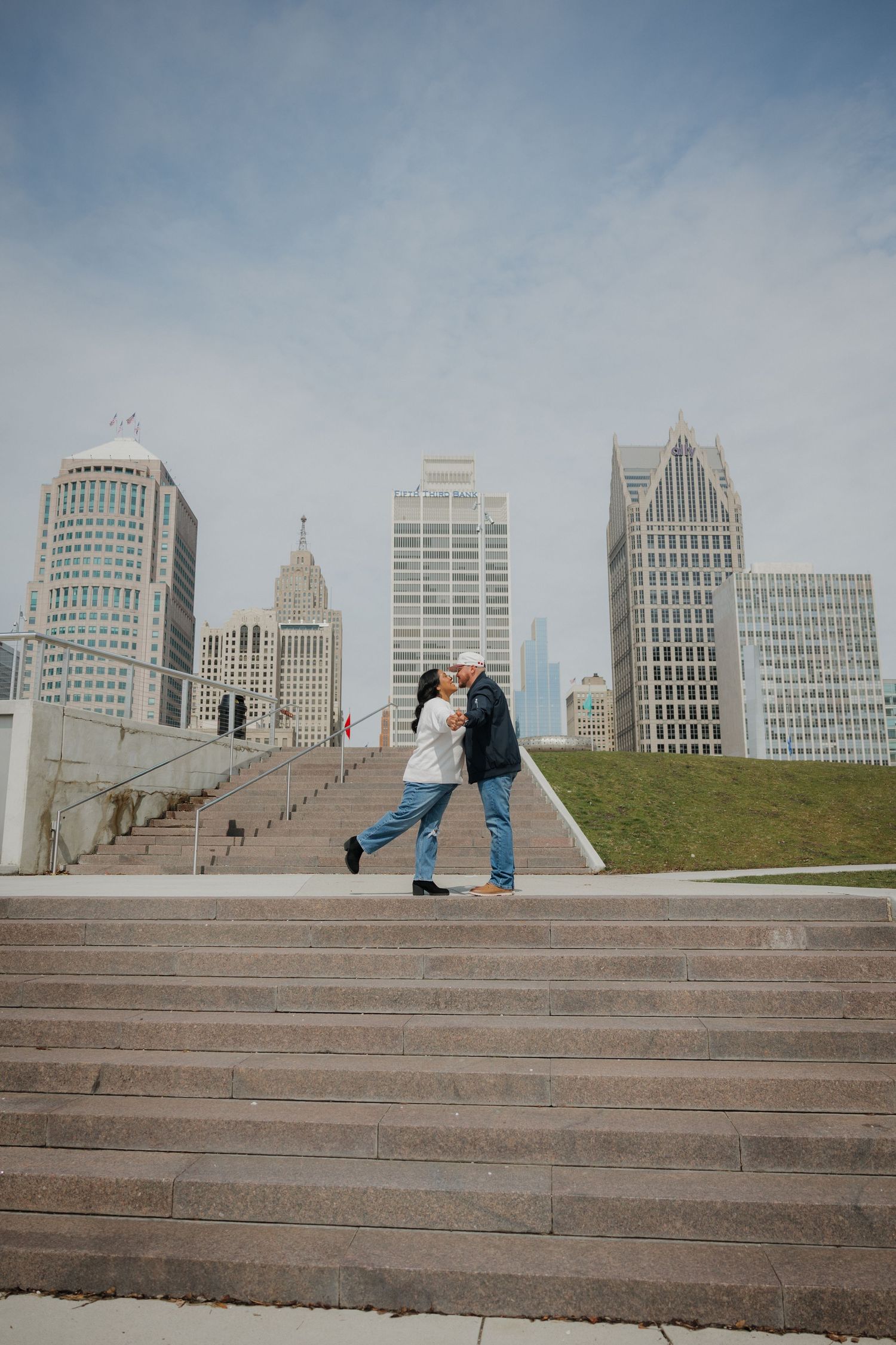 Couple embracing on steps with Detroit city skyline in background on a cloudy day.