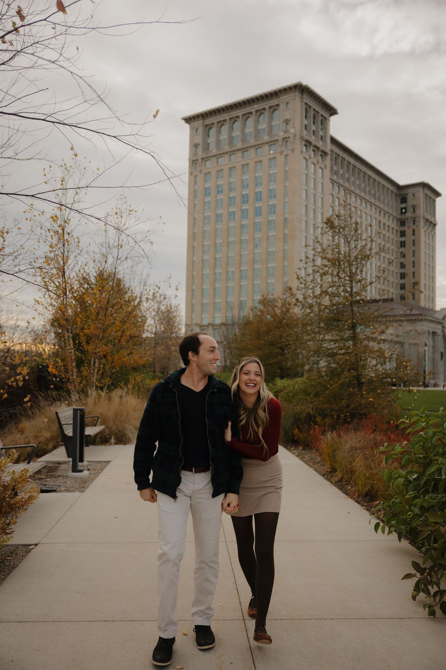 Couple walking and laughing on a pathway with autumn foliage and a tall historic building in the background.