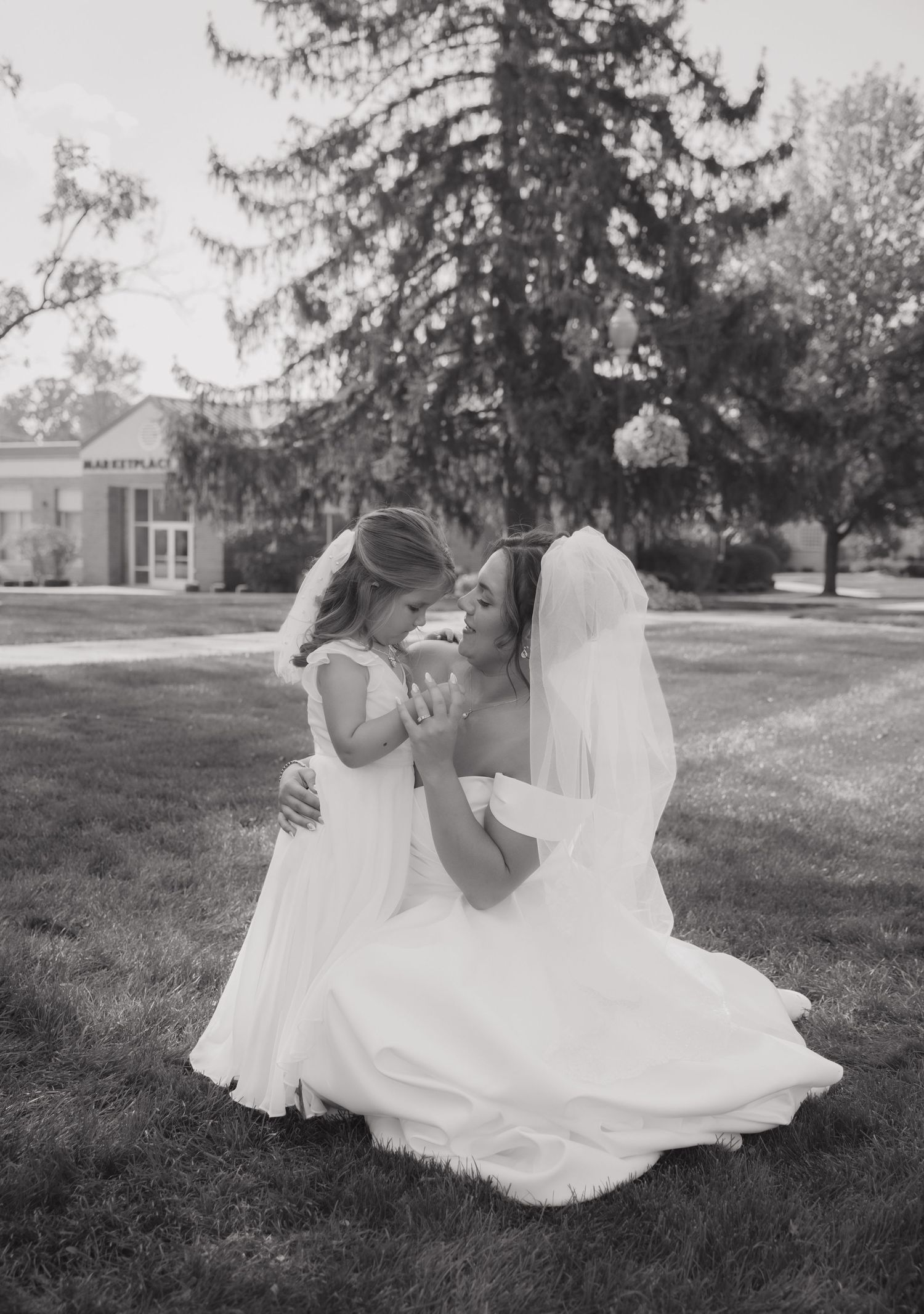 Black and white photo of a bride in a veil kneeling on grass, sharing a tender moment with a young flower girl in white dress.