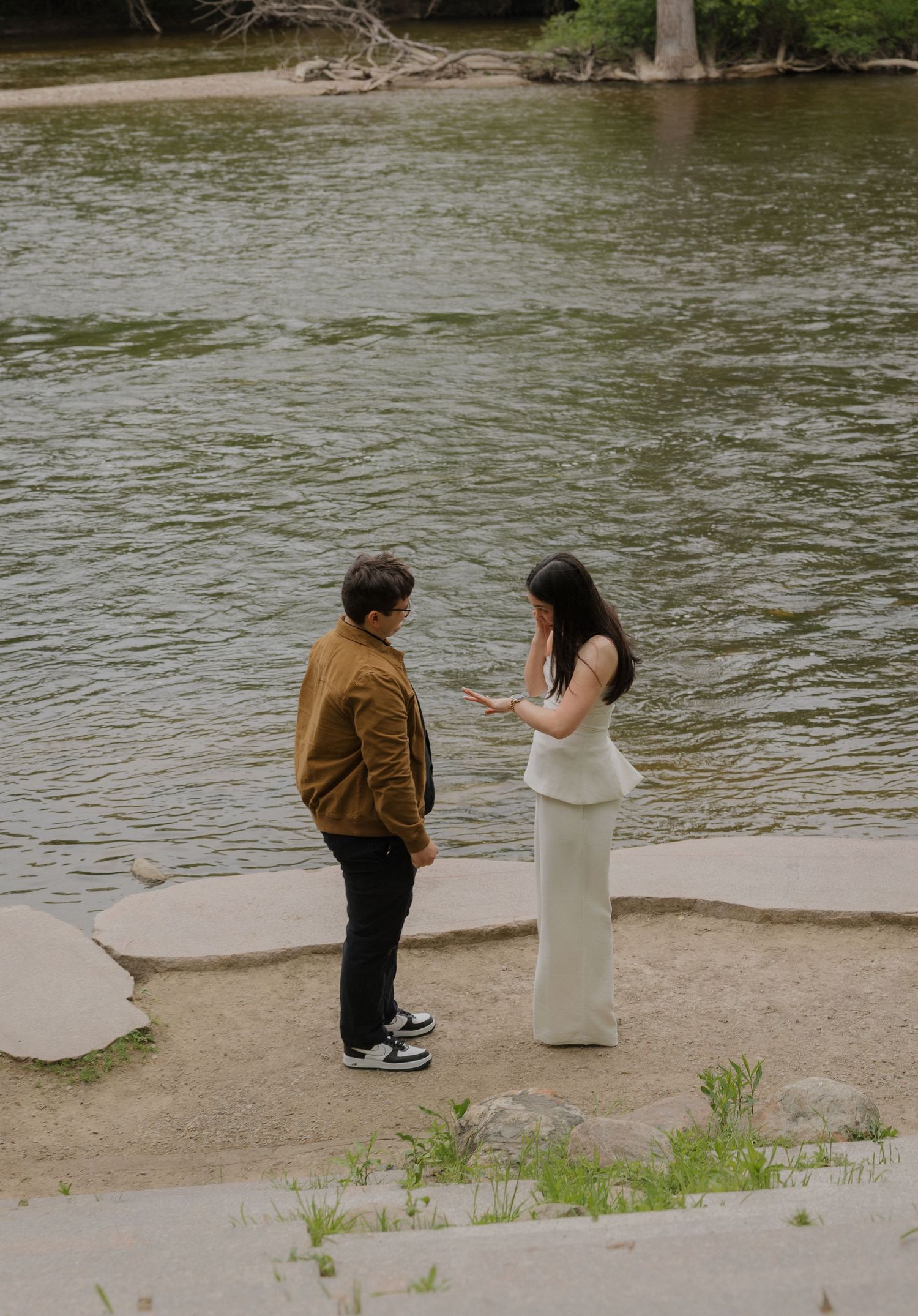 Couple exchanging vows by a serene river, woman in white dress holding notes, man in brown jacket on sandy shore.