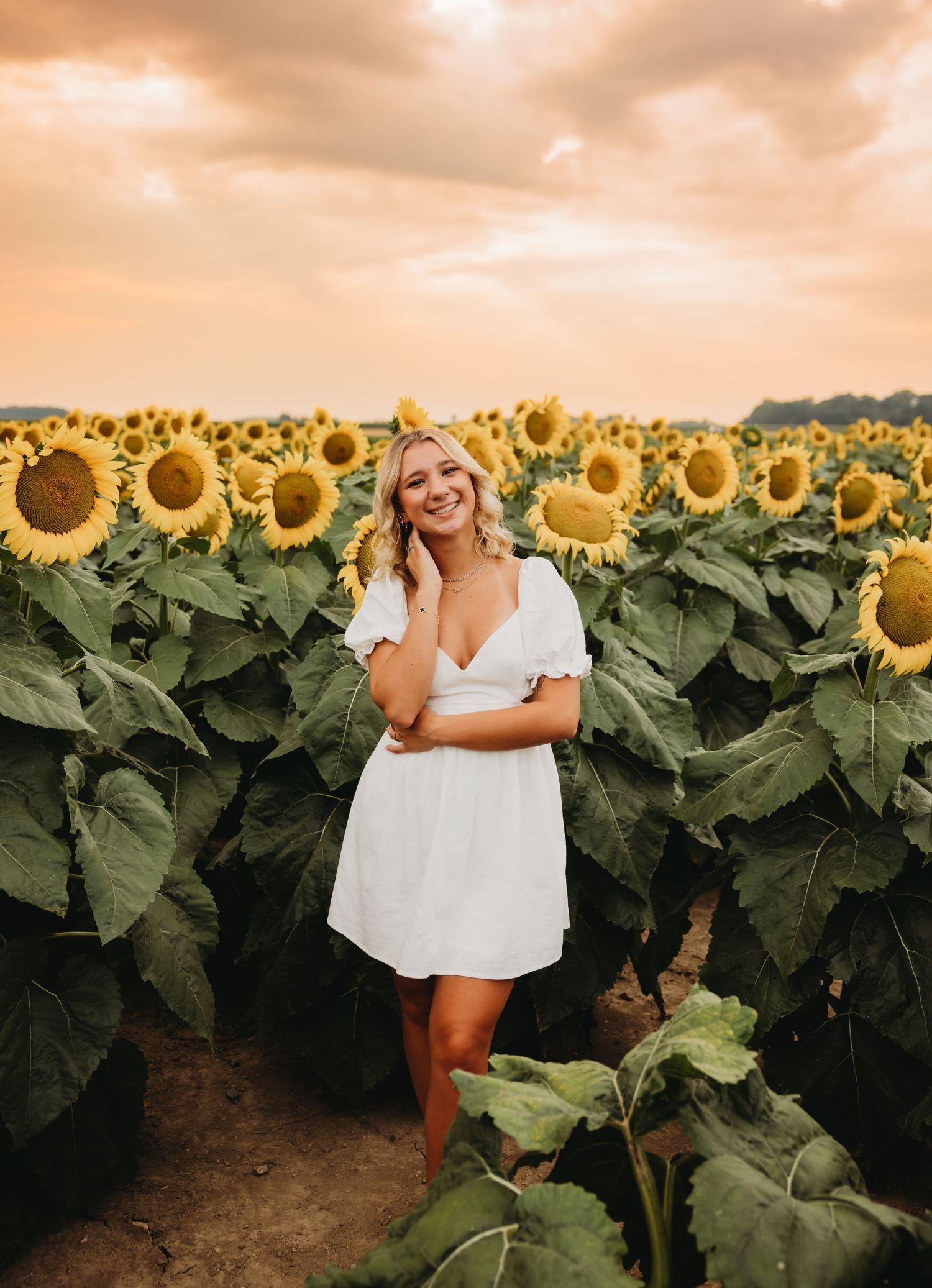 A smiling blonde woman in a white dress poses in a vibrant sunflower field at golden hour sunset.