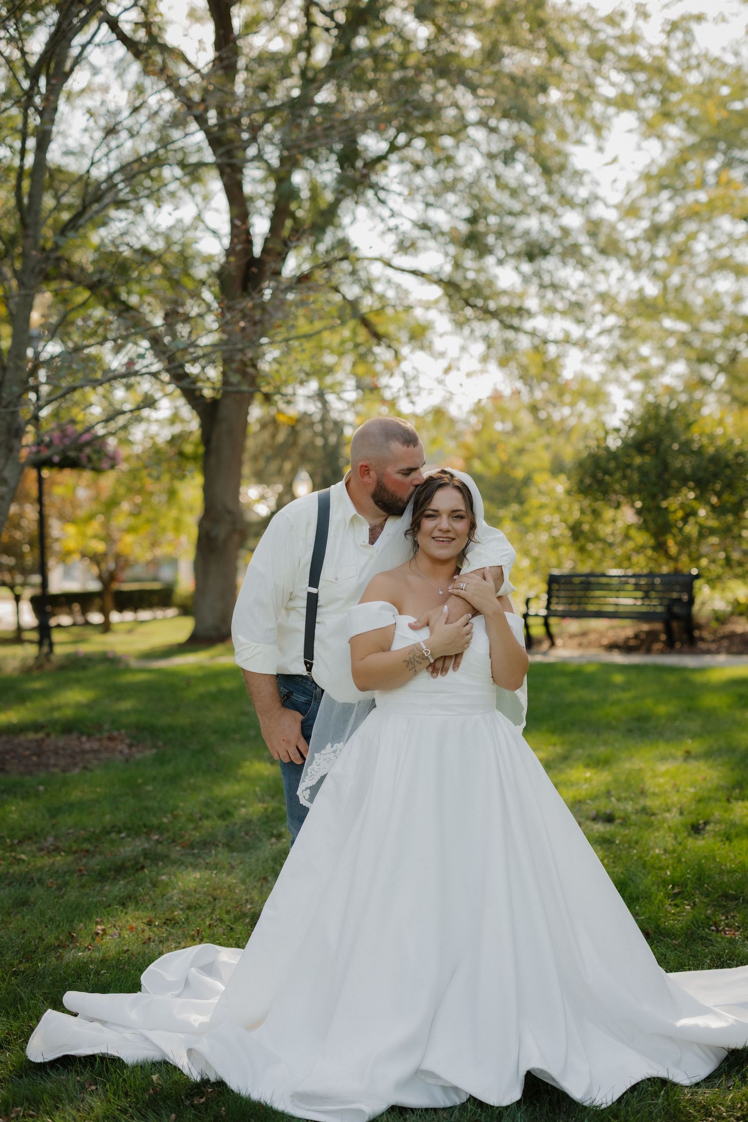 A bride in a white off-shoulder gown and groom in suspenders share a tender moment in a lush green park.