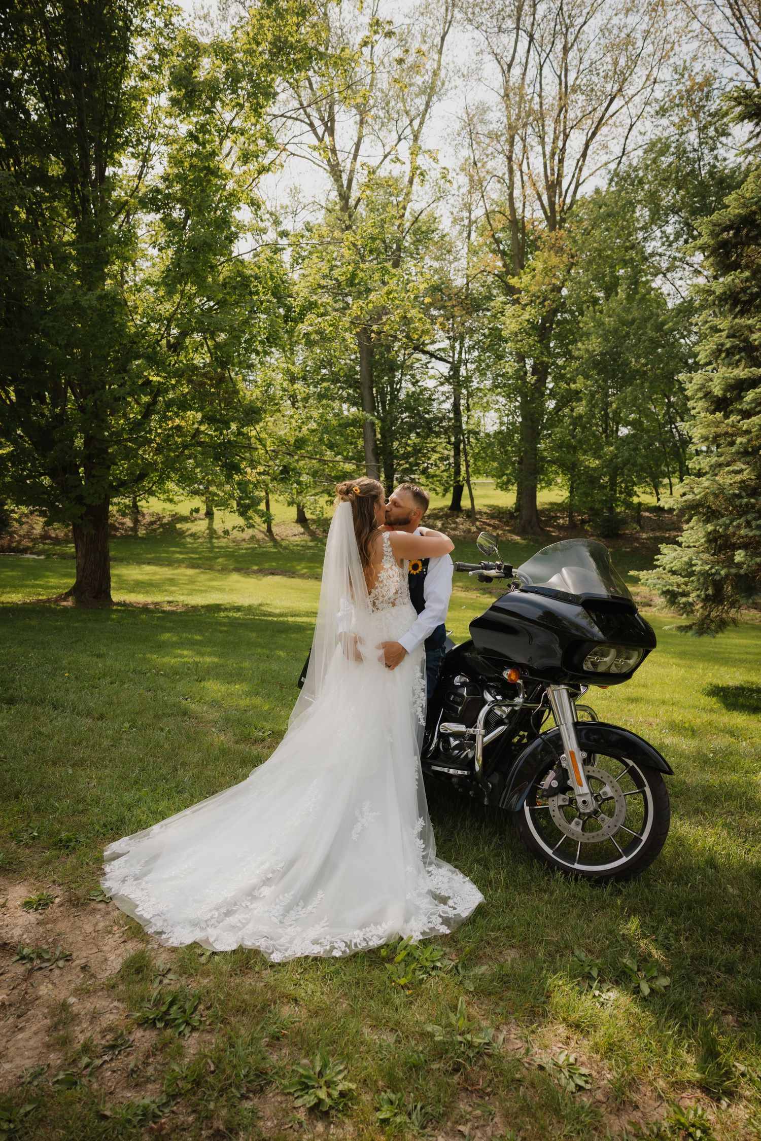 Bride and groom embrace near a Harley-Davidson motorcycle in a lush green outdoor wedding setting.