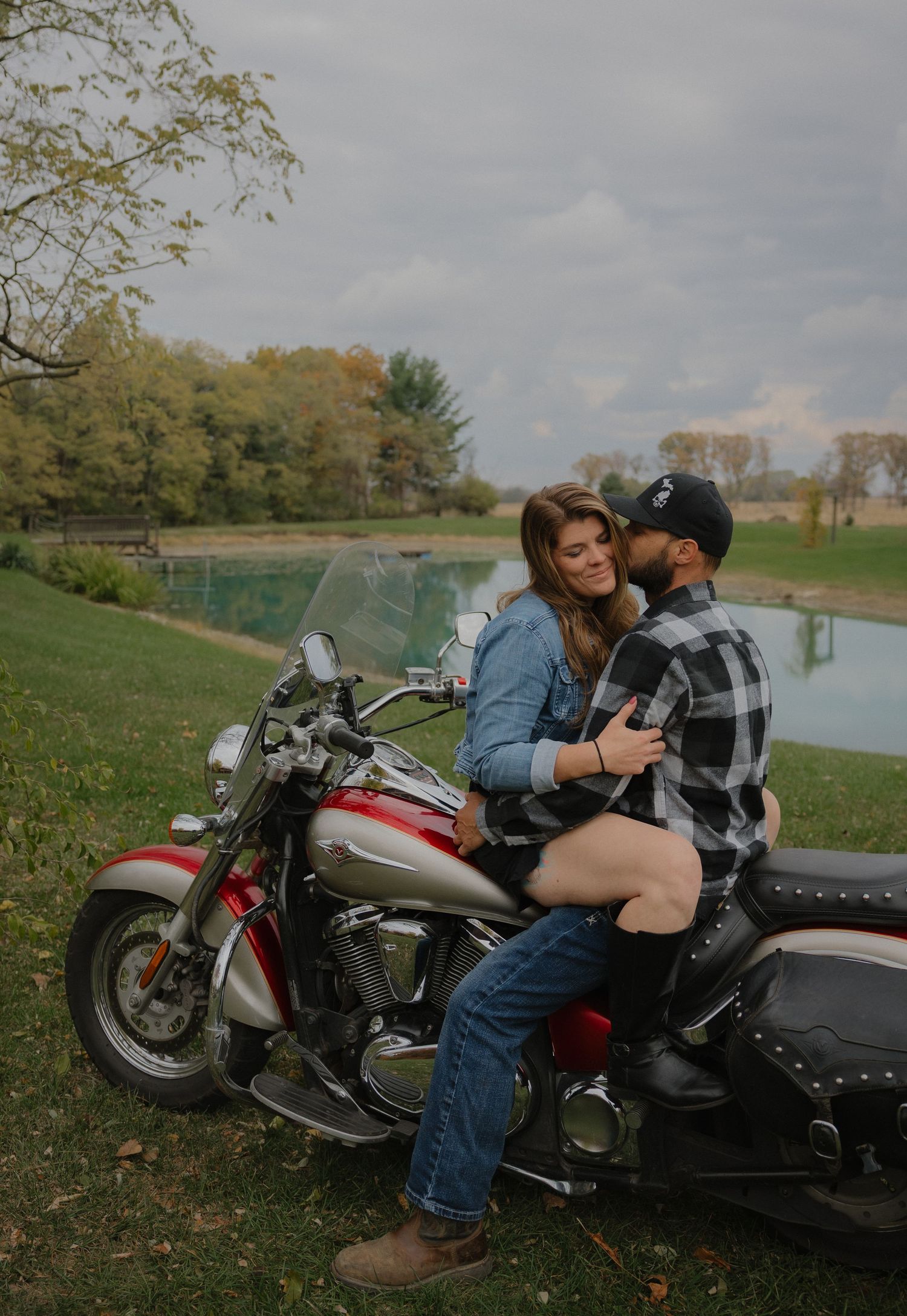 A couple shares an intimate moment next to a vintage motorcycle in a scenic autumn field with a lake in the background.