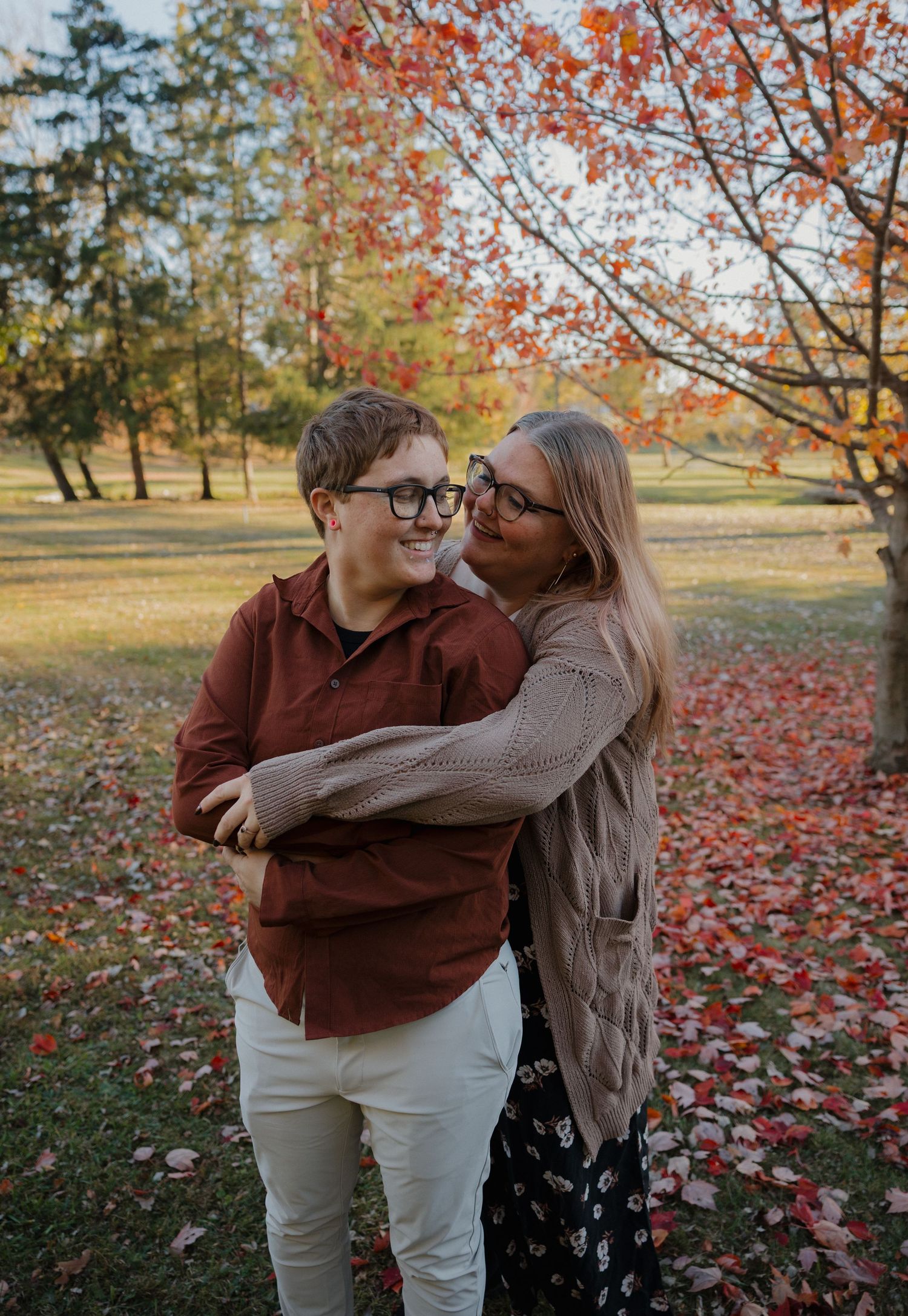 A mother and son share a warm embrace outdoors during autumn, surrounded by vibrant red and orange foliage.