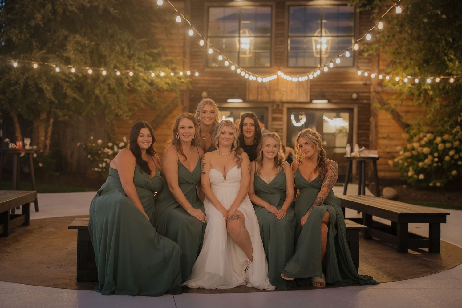Wedding party poses together under twinkling string lights at a rustic outdoor venue.