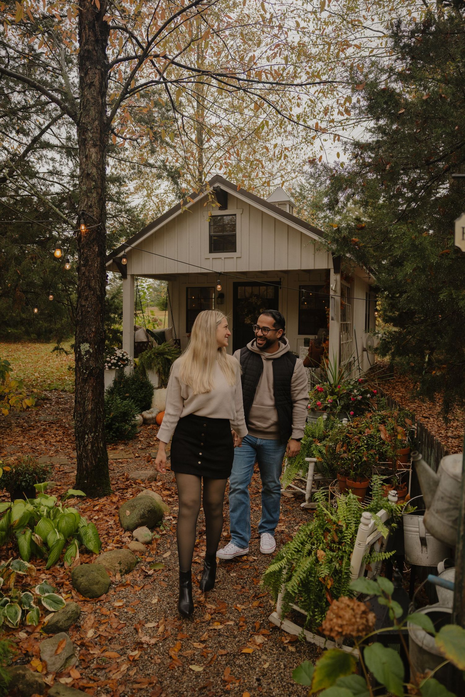 Cozy autumn scene with fallen leaves surrounding a rustic cottage and garden with lush green plants.