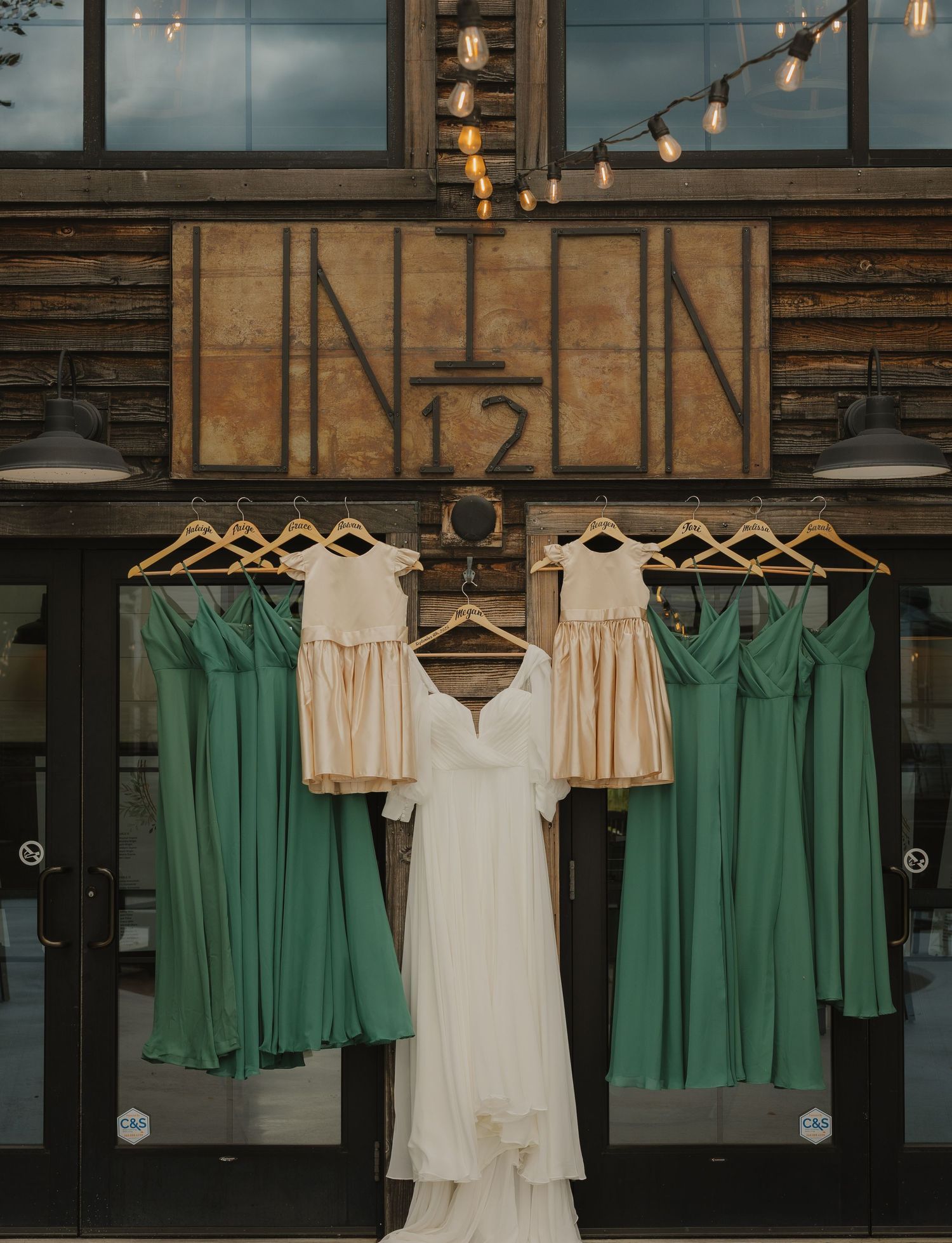 Wedding dresses and mint green bridesmaid dresses hanging on display in rustic venue with string lights.