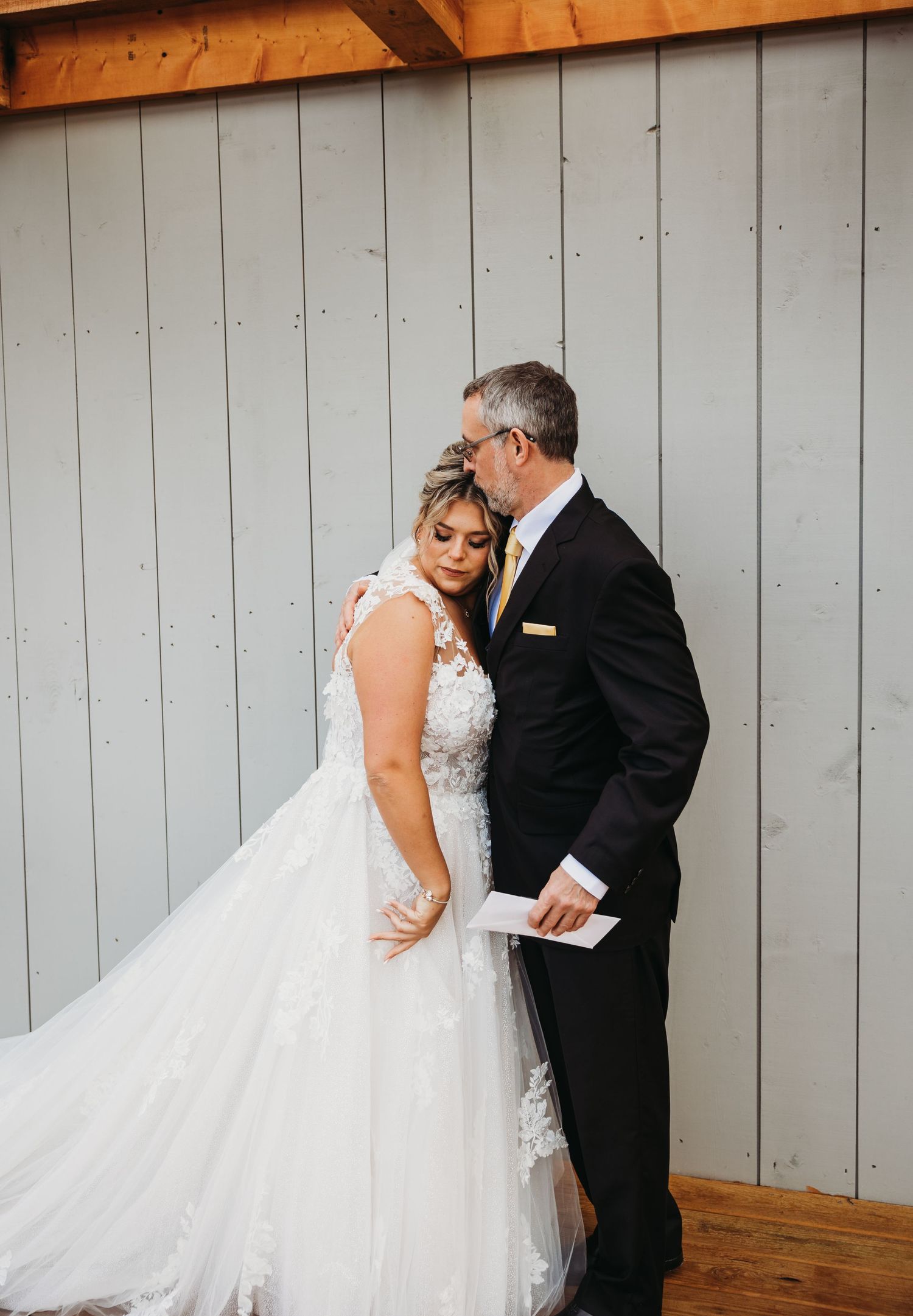 Intimate moment between bride in lace gown and person in suit against grey wall.