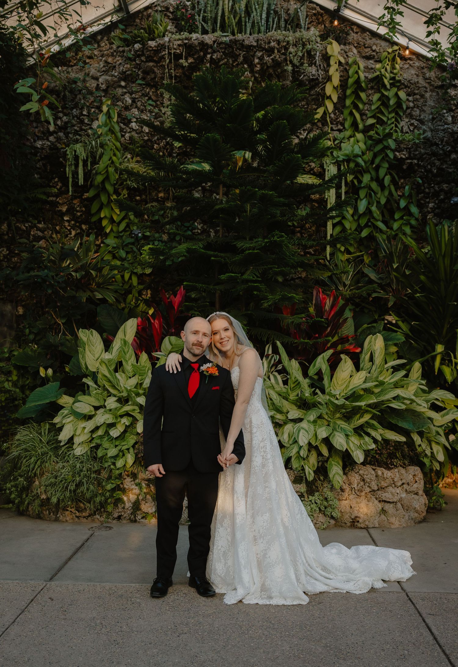 Bride and groom posing together in a lush greenhouse garden, bride in white lace gown, groom in black suit with red tie.