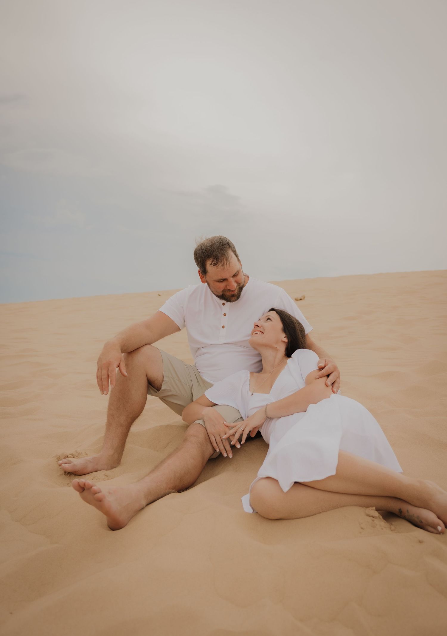 Couple sitting together on sandy desert dunes, woman in white dress leaning on man in casual attire.