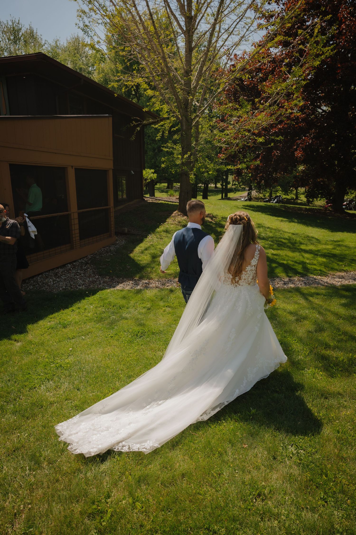 Bride in white gown with veil and groom in navy suit walk across green lawn toward rustic building on sunny wedding day.