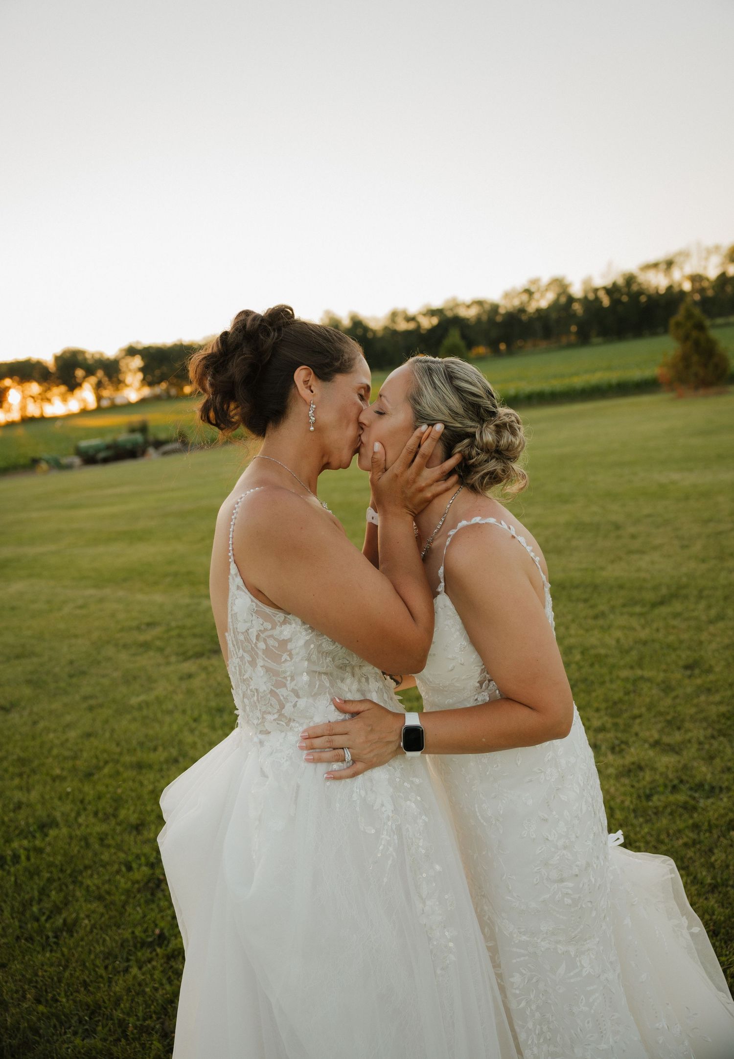 Two brides in white lace gowns share a romantic kiss at golden hour on a lush green lawn.