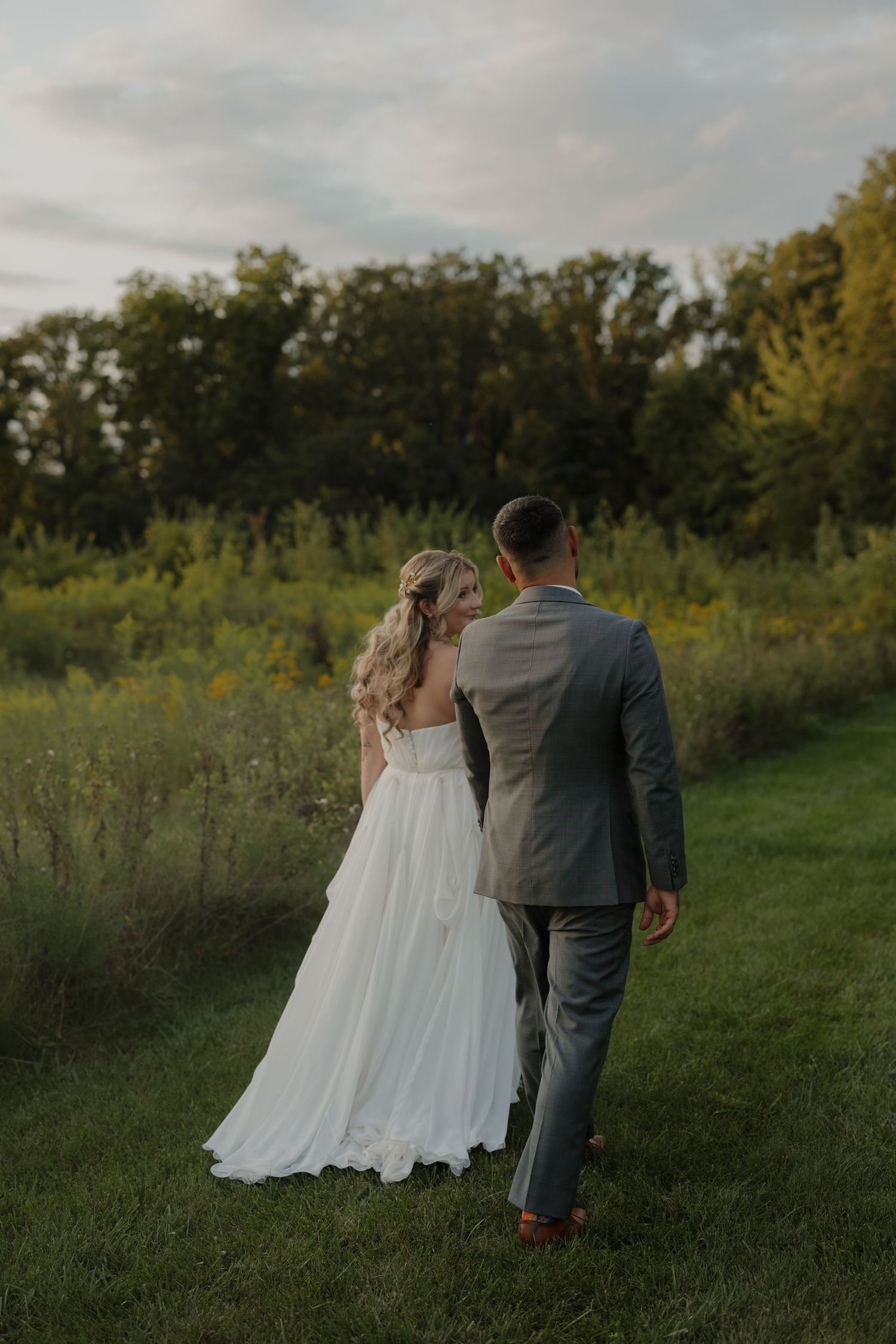 A couple in wedding attire walks hand in hand through a lush green field at sunset.
