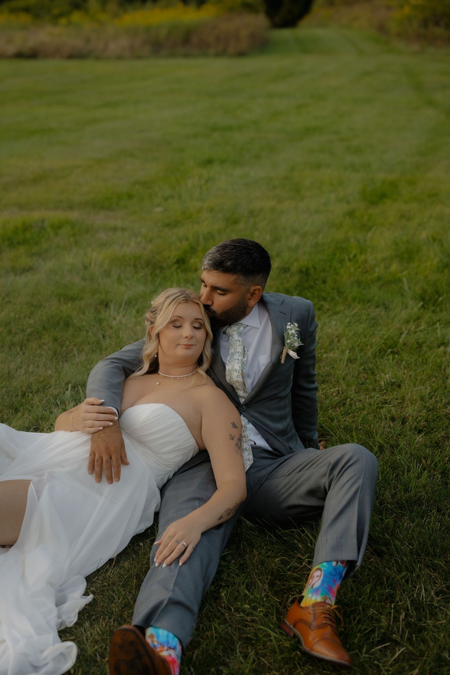 Couple in wedding attire relaxes on grassy field during outdoor photo session.