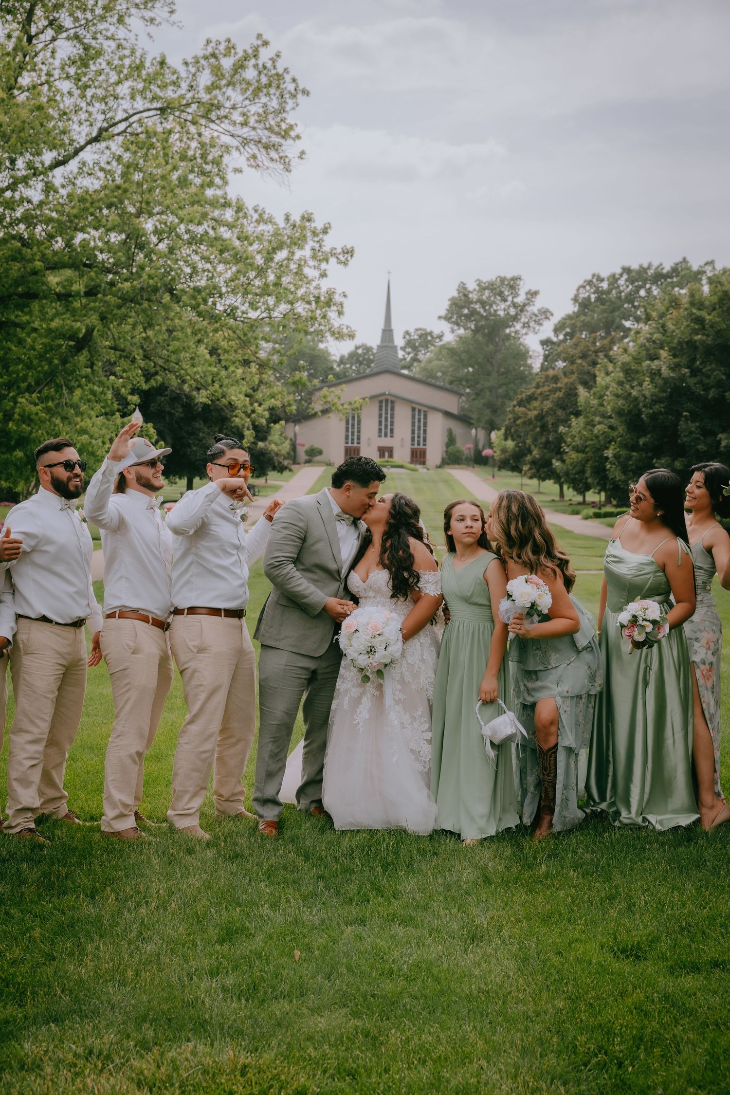 Bridal party with couple poses in casual line formation on grassy lawn wearing sage green and light colored attire.