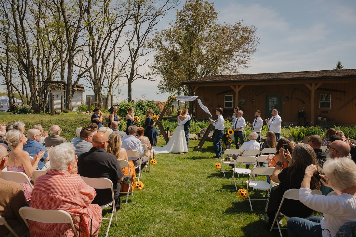 Outdoor wedding ceremony on green lawn with guests seated in white chairs near a brick building on a sunny day.