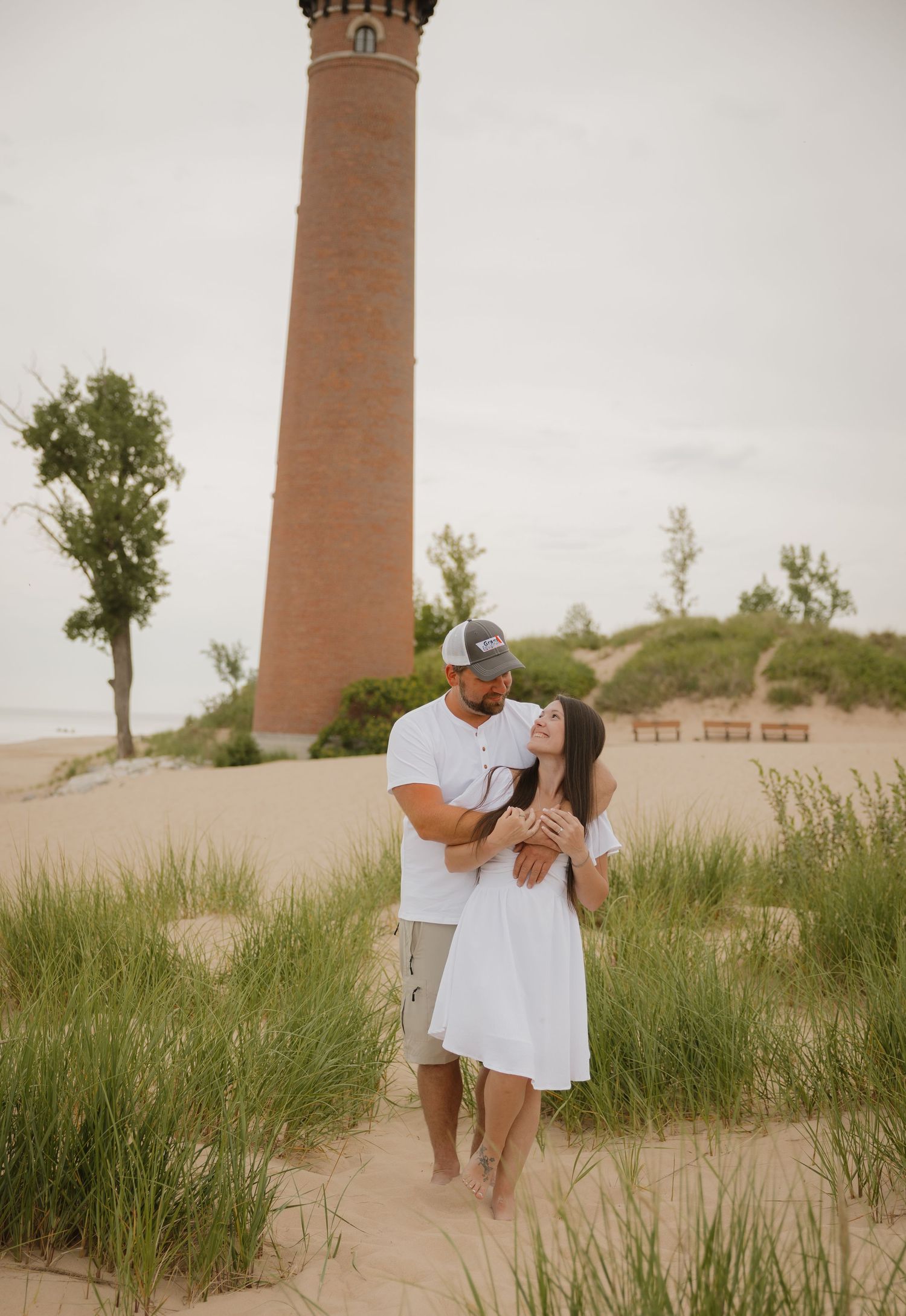 Couple embracing on sandy beach dunes near a tall brick lighthouse surrounded by green trees.