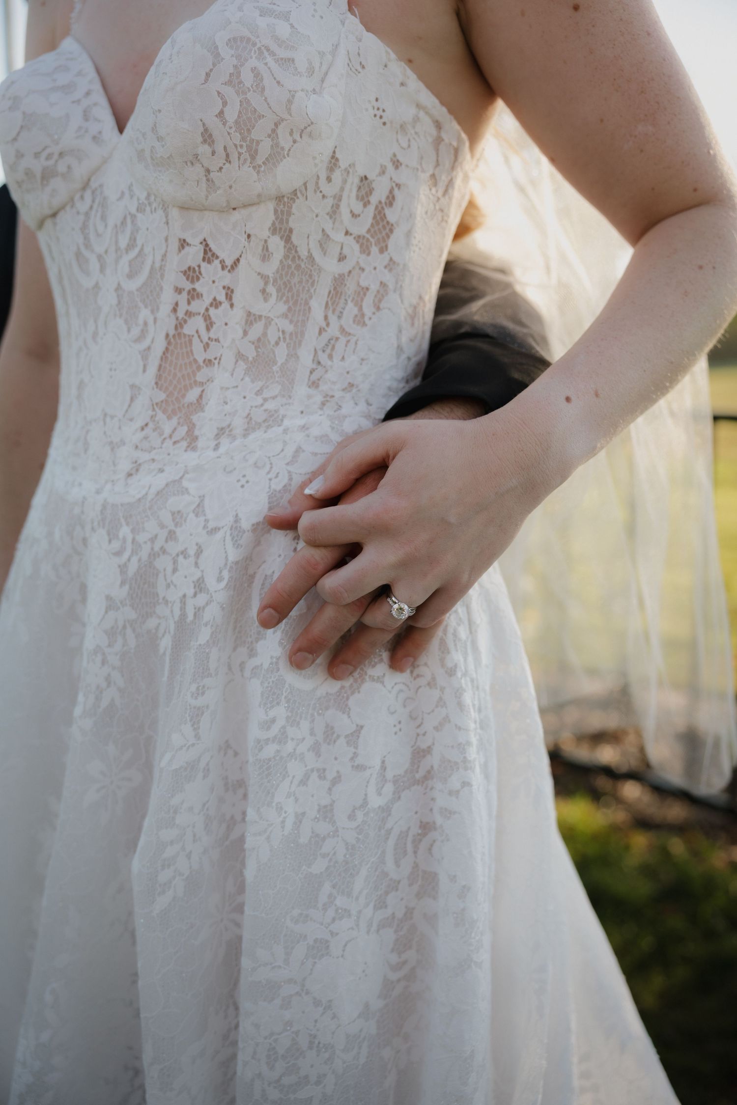 Close-up of a bride in a white lace wedding gown holding hands with groom, showcasing a diamond engagement ring.